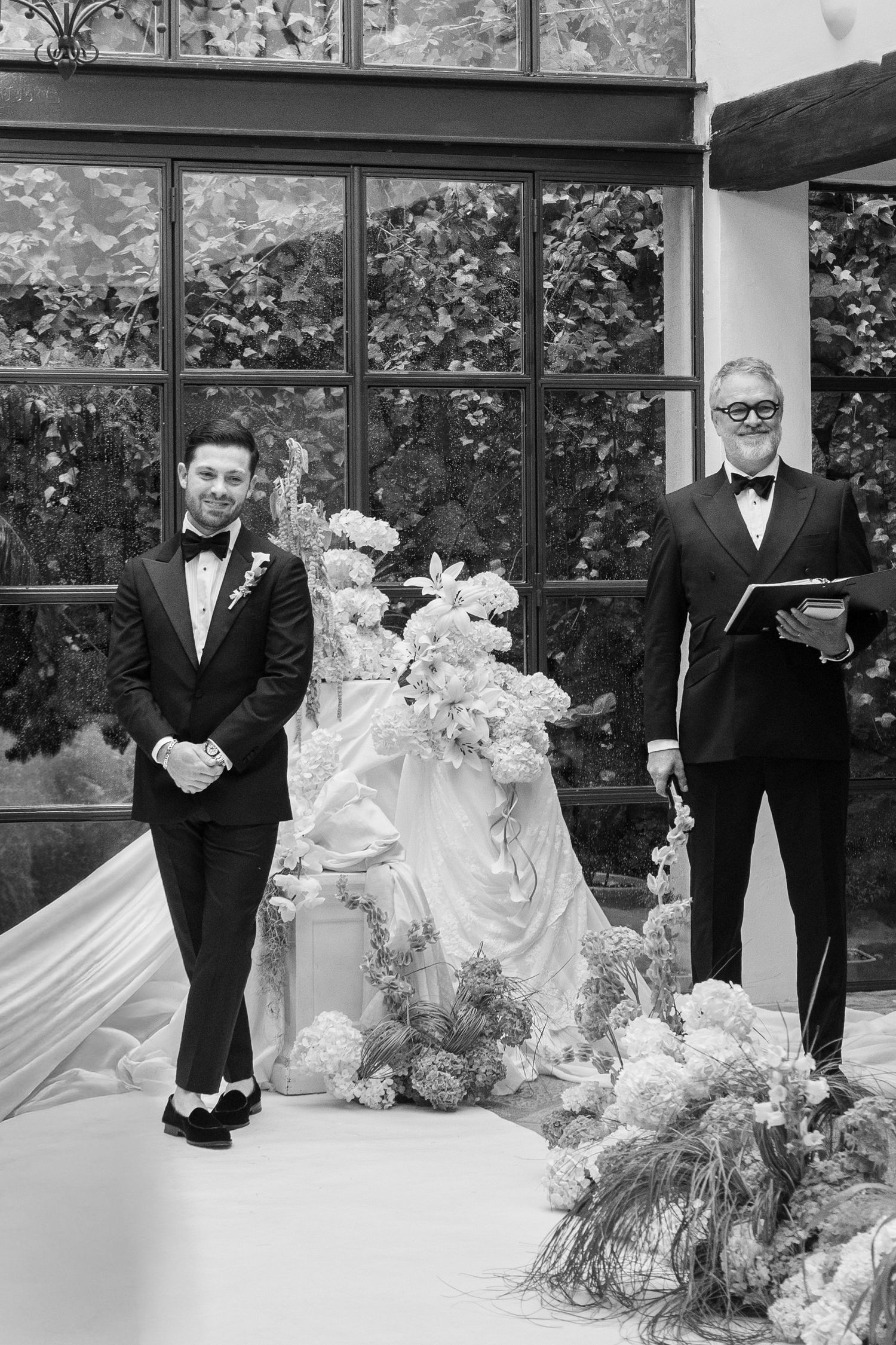 Wedding party in formal attire arranged along white floral aisle in greenhouse setting.