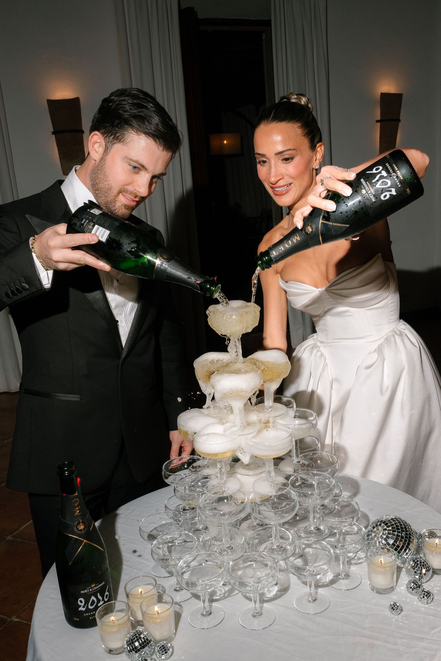 A couple pours champagne into a tower of coupe glasses creating an elegant champagne fountain at a formal event.