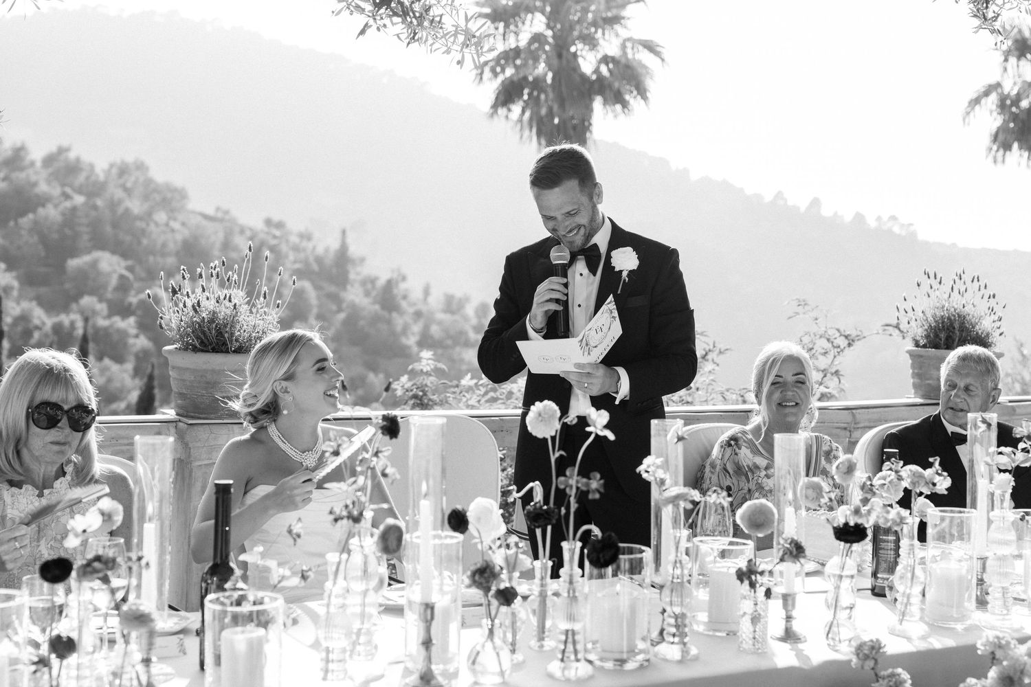 Wedding guests seated at elegant tables with floral arrangements on a sunny terrace.