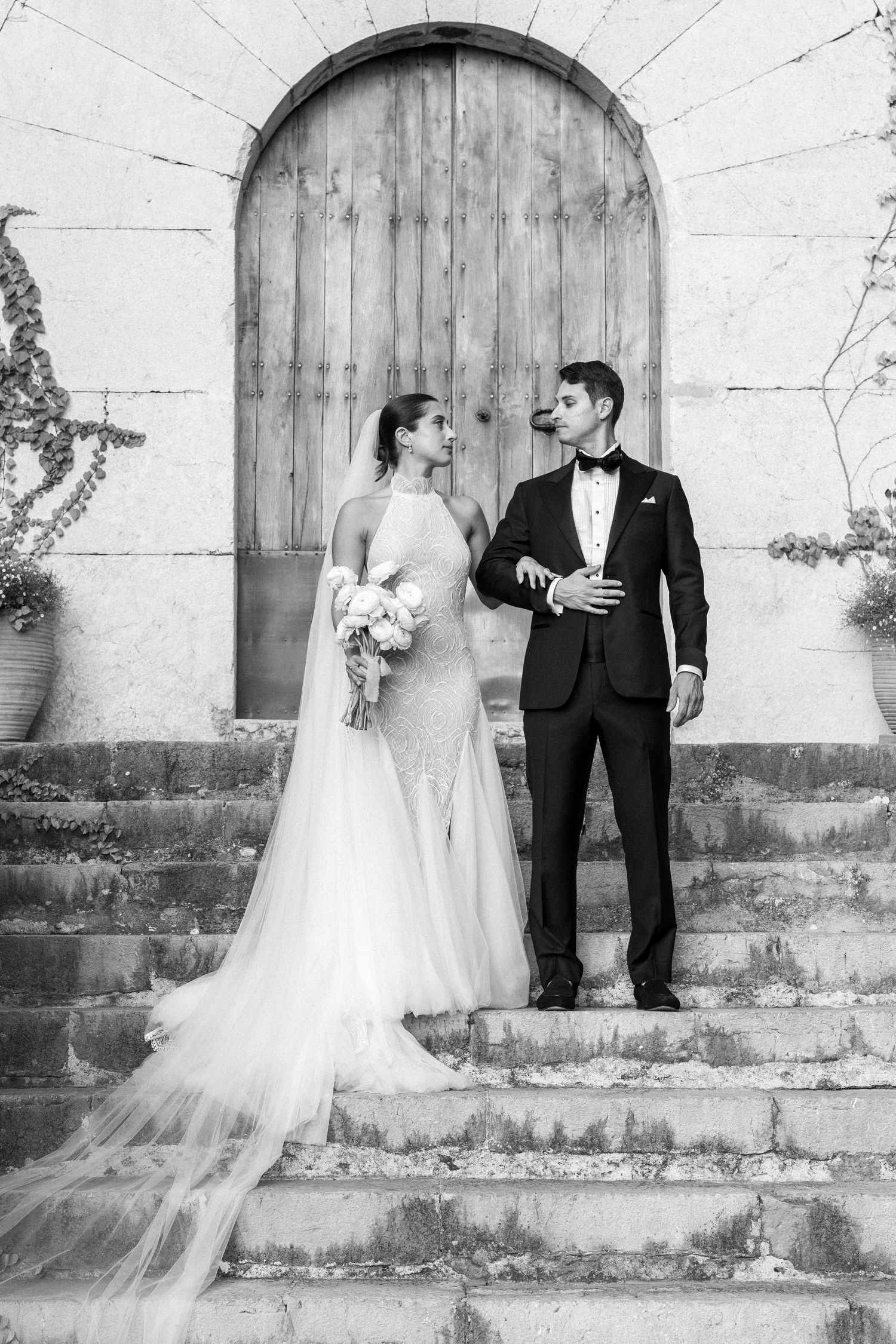 A bride and groom pose elegantly on stone steps in front of a rustic wooden door in black and white.