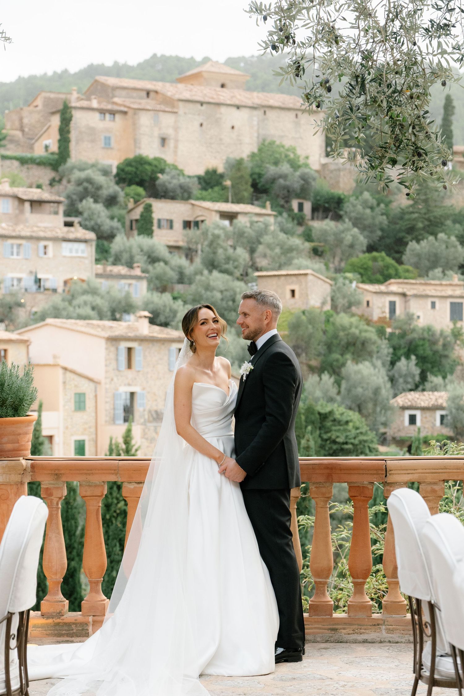 A couple in wedding attire embraces on a terrace overlooking a Mediterranean hillside village with stone buildings and olive trees.