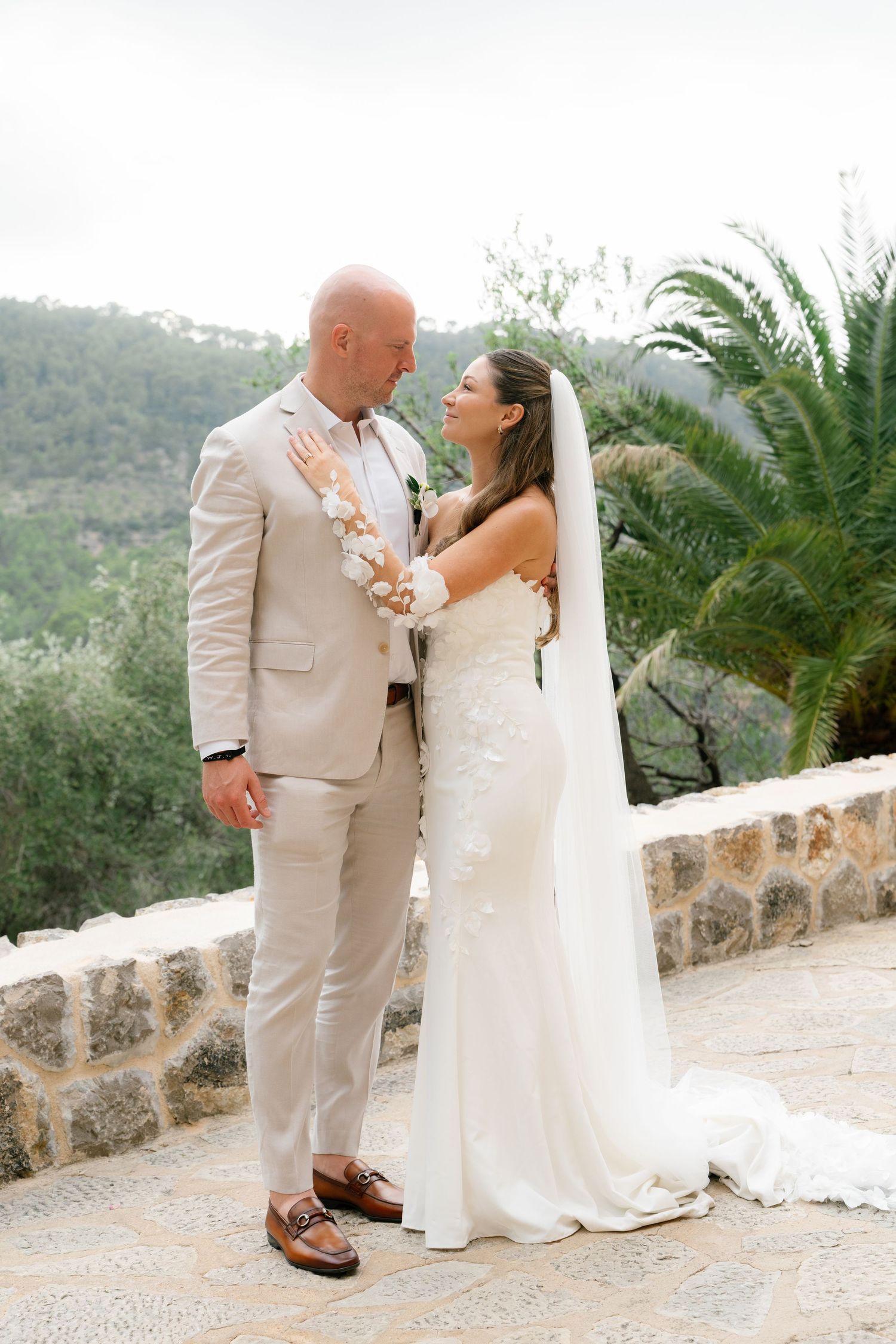 Newlyweds walk together on stone terrace with palm trees and mountain views.