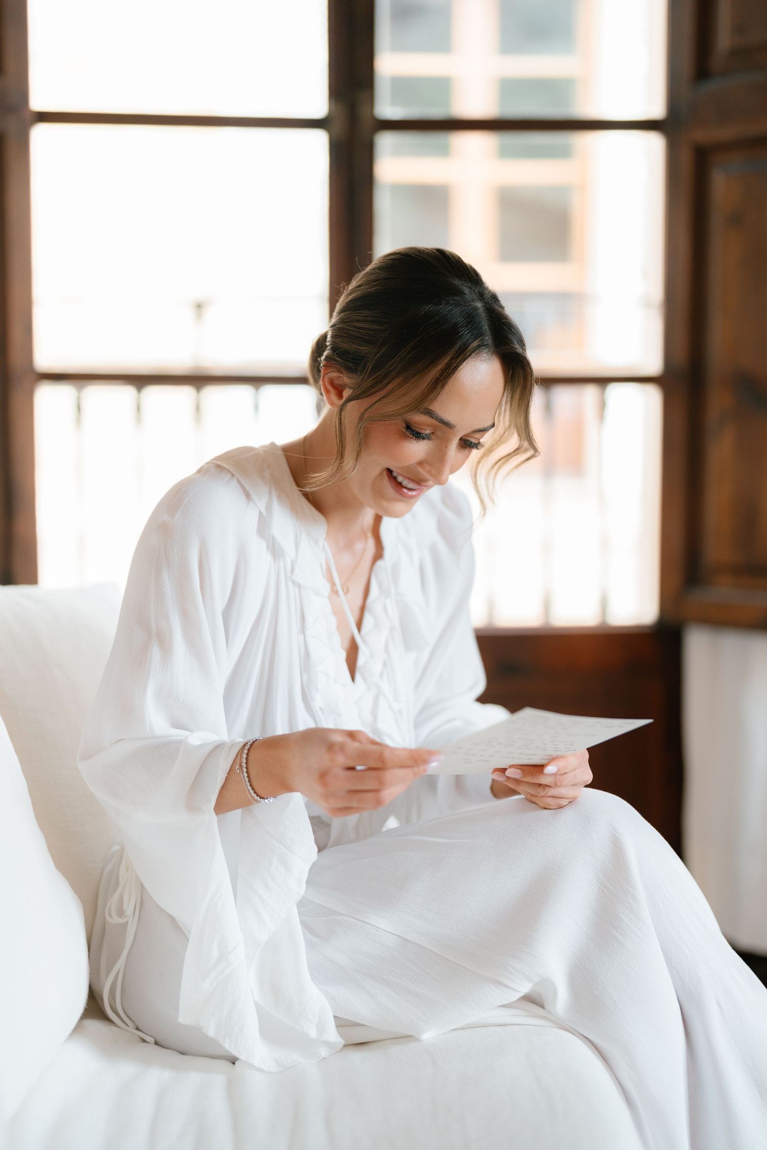 Bride in white robe reading letter while sitting by window in morning light.
