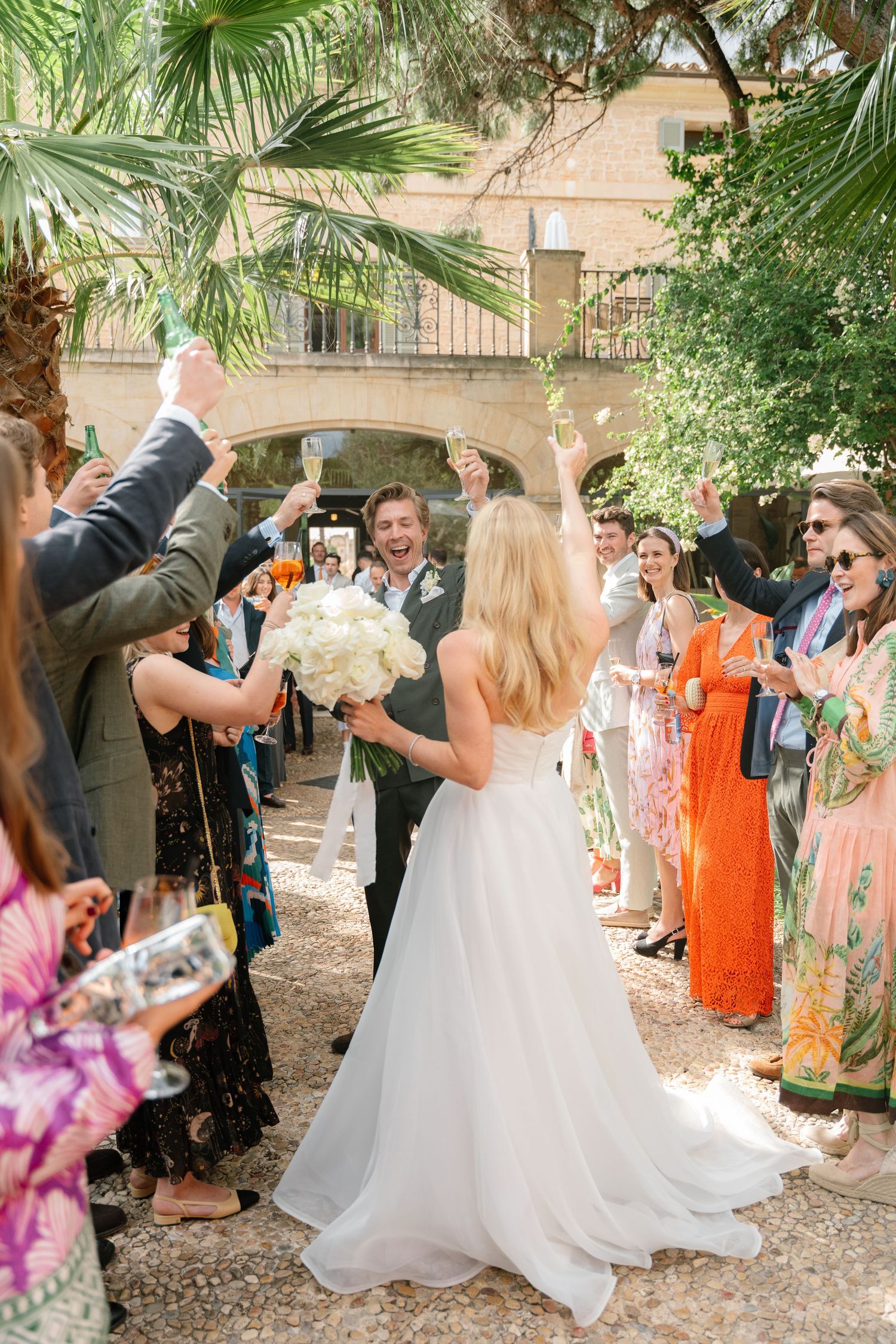 Wedding guests gather under palm trees and greenery during an outdoor celebration.
