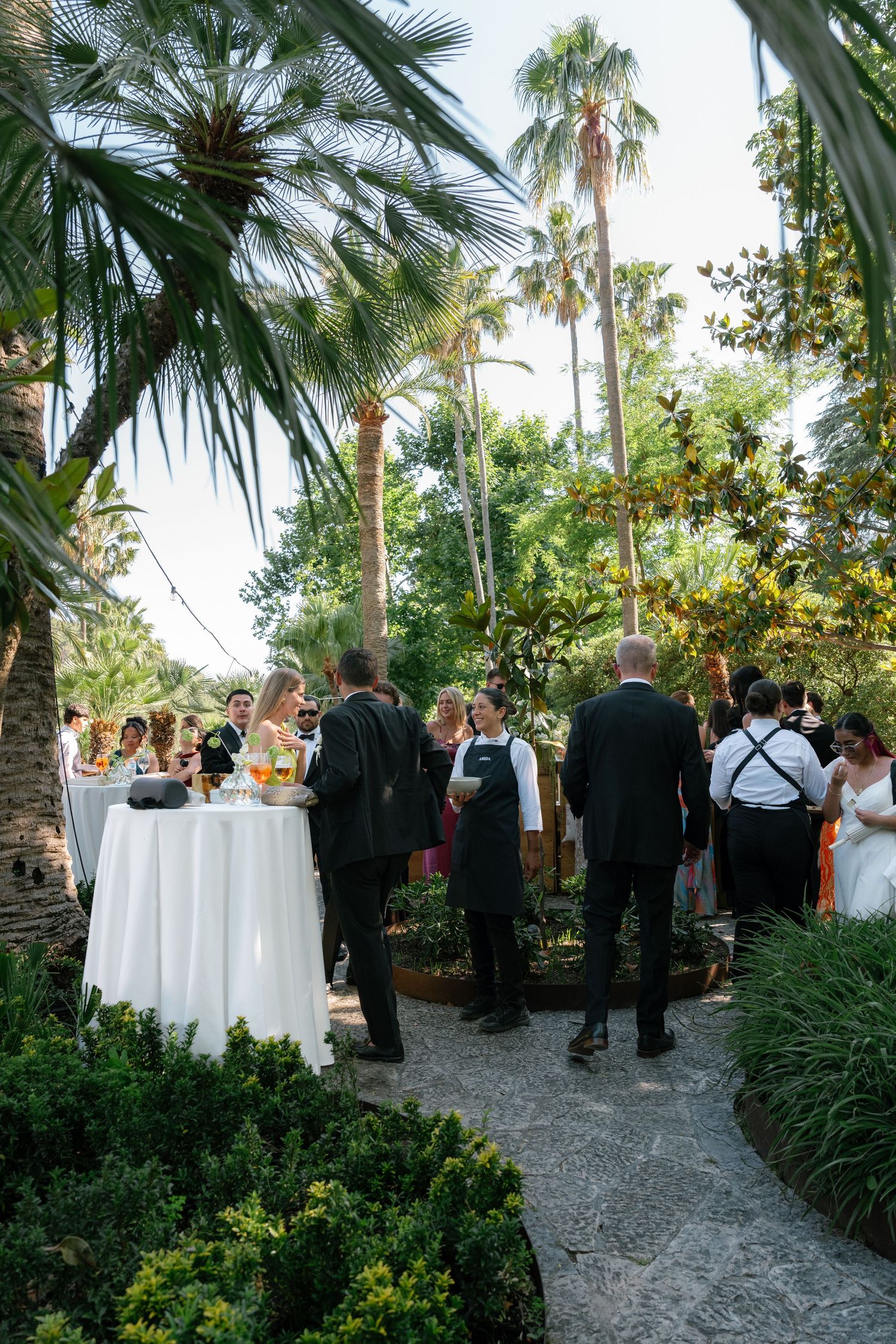 Elegant outdoor garden cocktail reception with guests mingling among palm trees and white-clothed high tables.