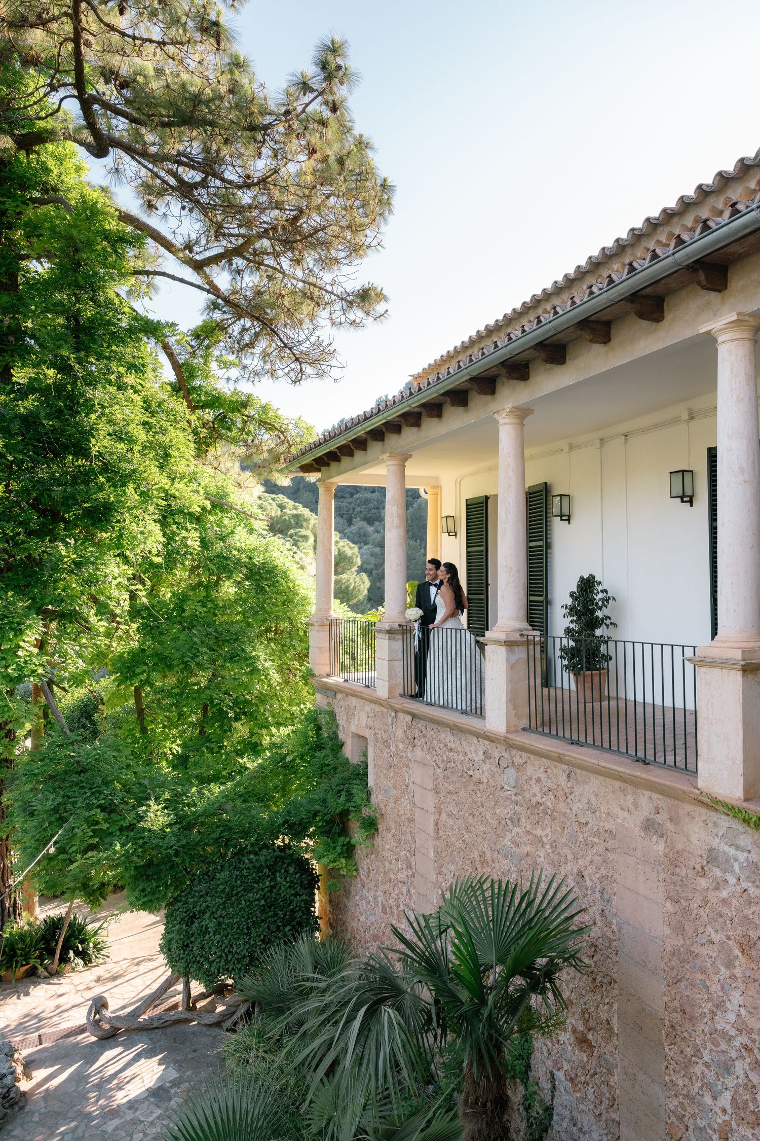 Couple embracing on elegant white villa balcony surrounded by lush green trees and Mediterranean gardens.