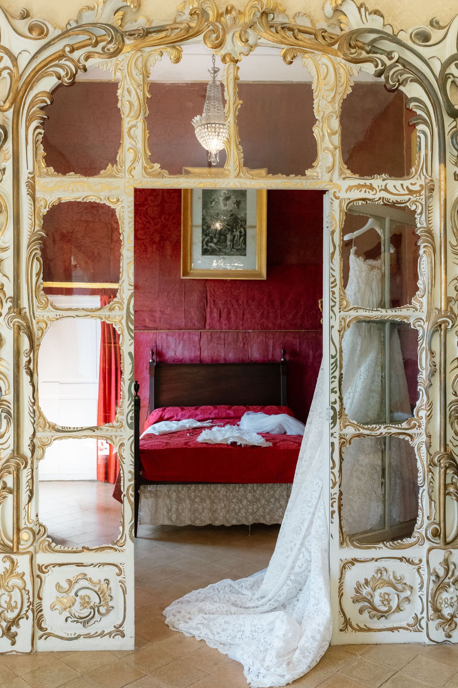 Ornate gold baroque doorway frames a red velvet bedroom with antique bed and hanging wedding dress.