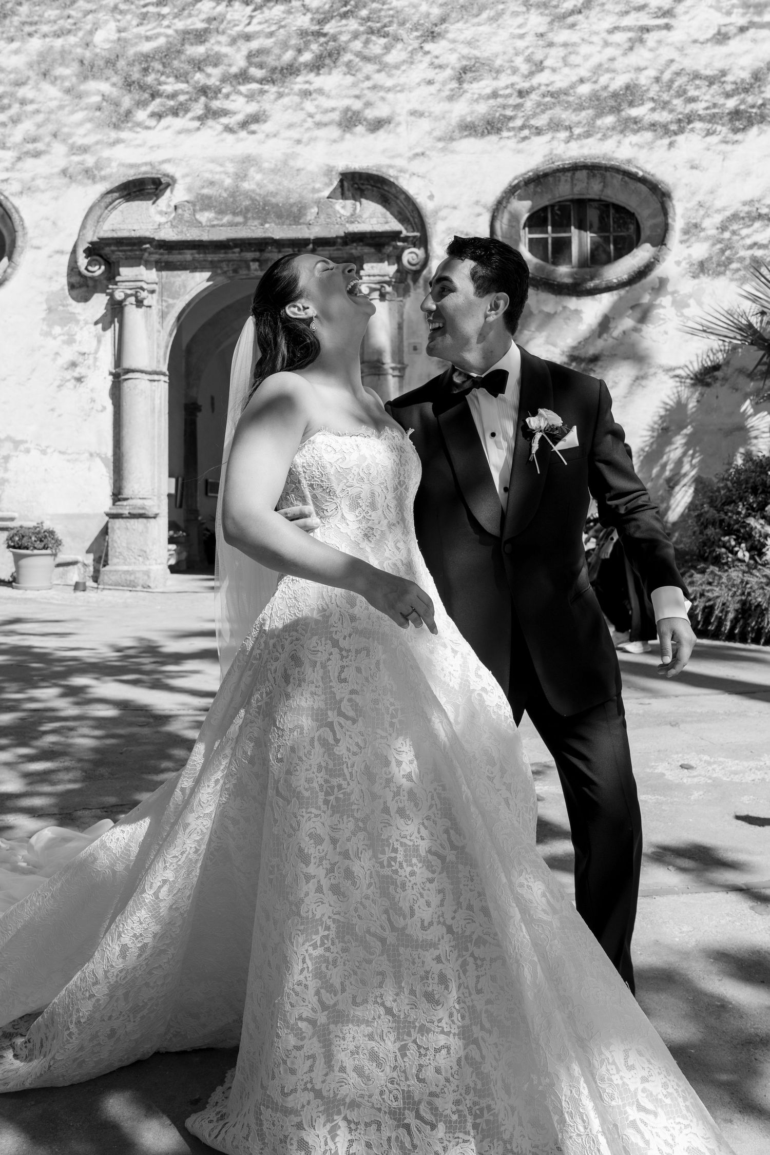 Black and white photo of a bride and groom sharing a kiss outside a historic stone building on their wedding day.