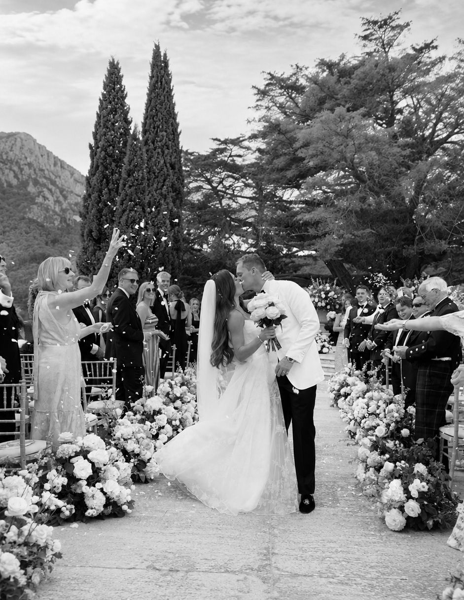 Black and white wedding photo of couple kissing at floral aisle, guests celebrating with confetti, trees and mountains behind.