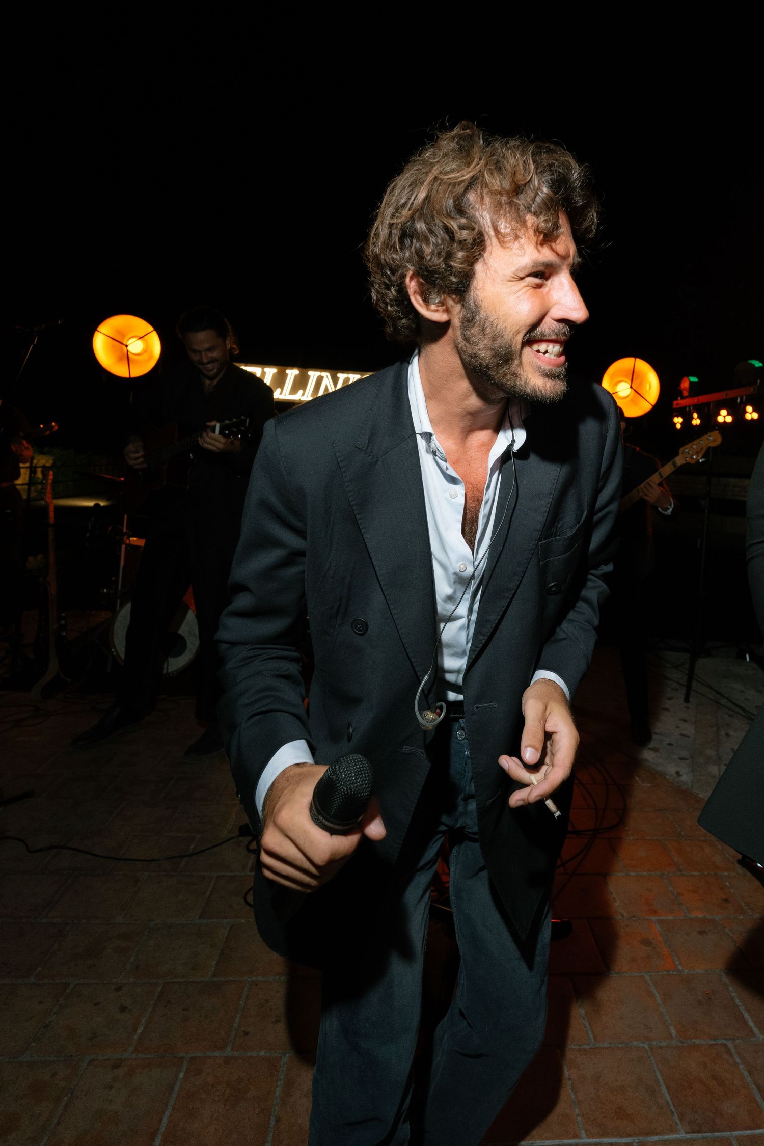 Man in dark suit holding microphone performing at night outdoor event with band and lights in background.