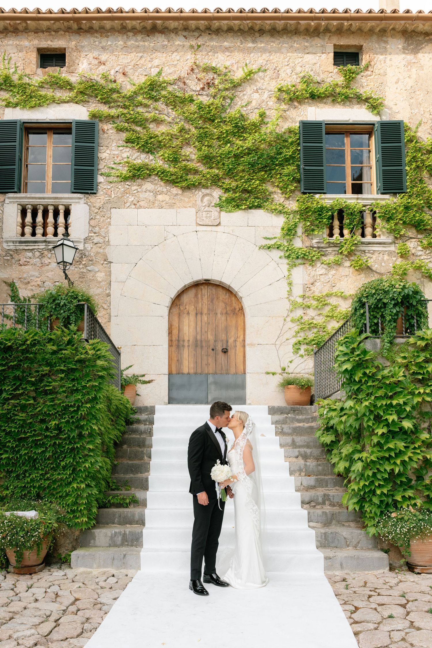 Bride and groom kissing on stone steps of an ivy-covered villa, bride holding white bouquet in elegant wedding attire.