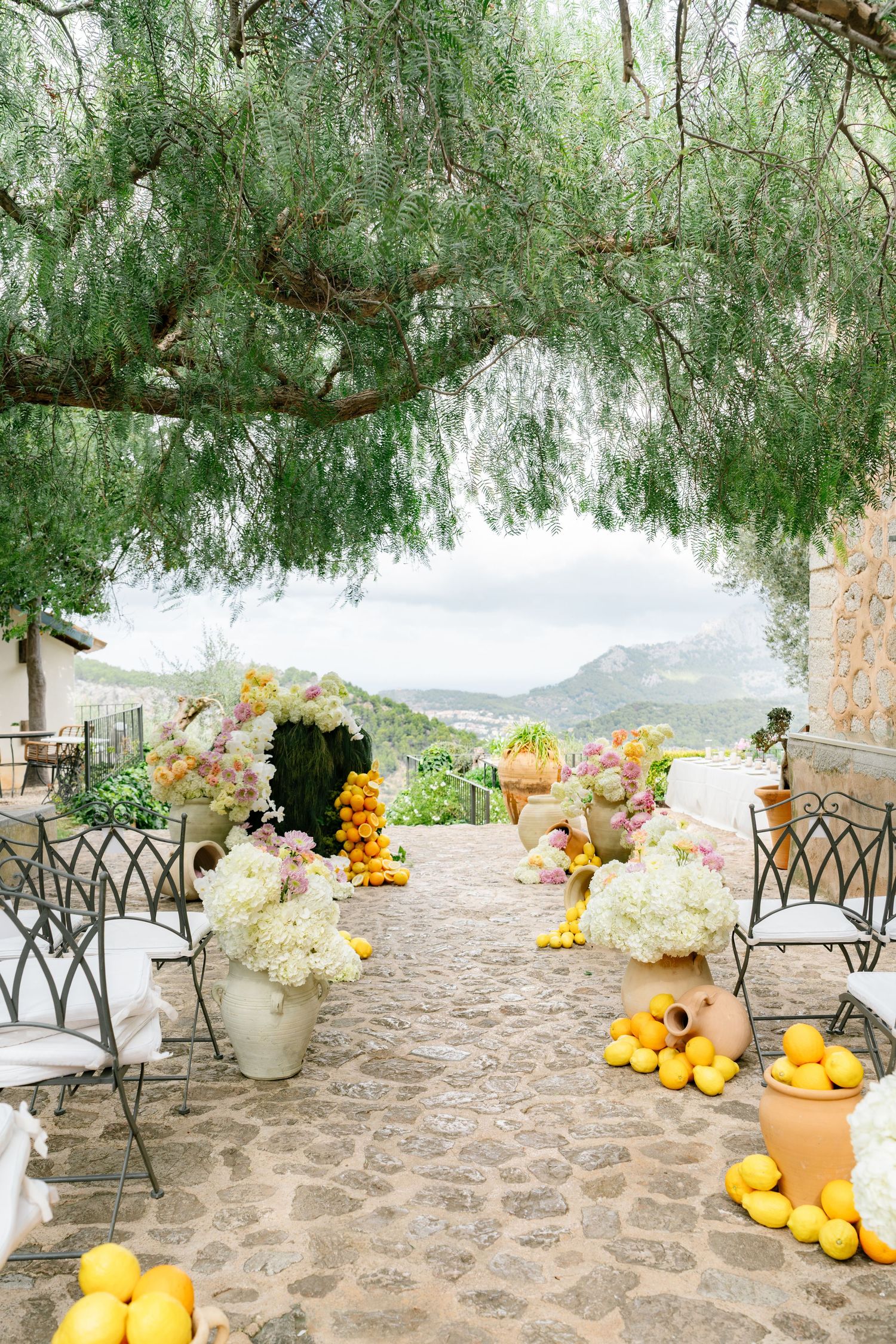 Outdoor wedding venue with stone patio decorated with white flowers, citrus fruits and willow trees overlooking mountains.