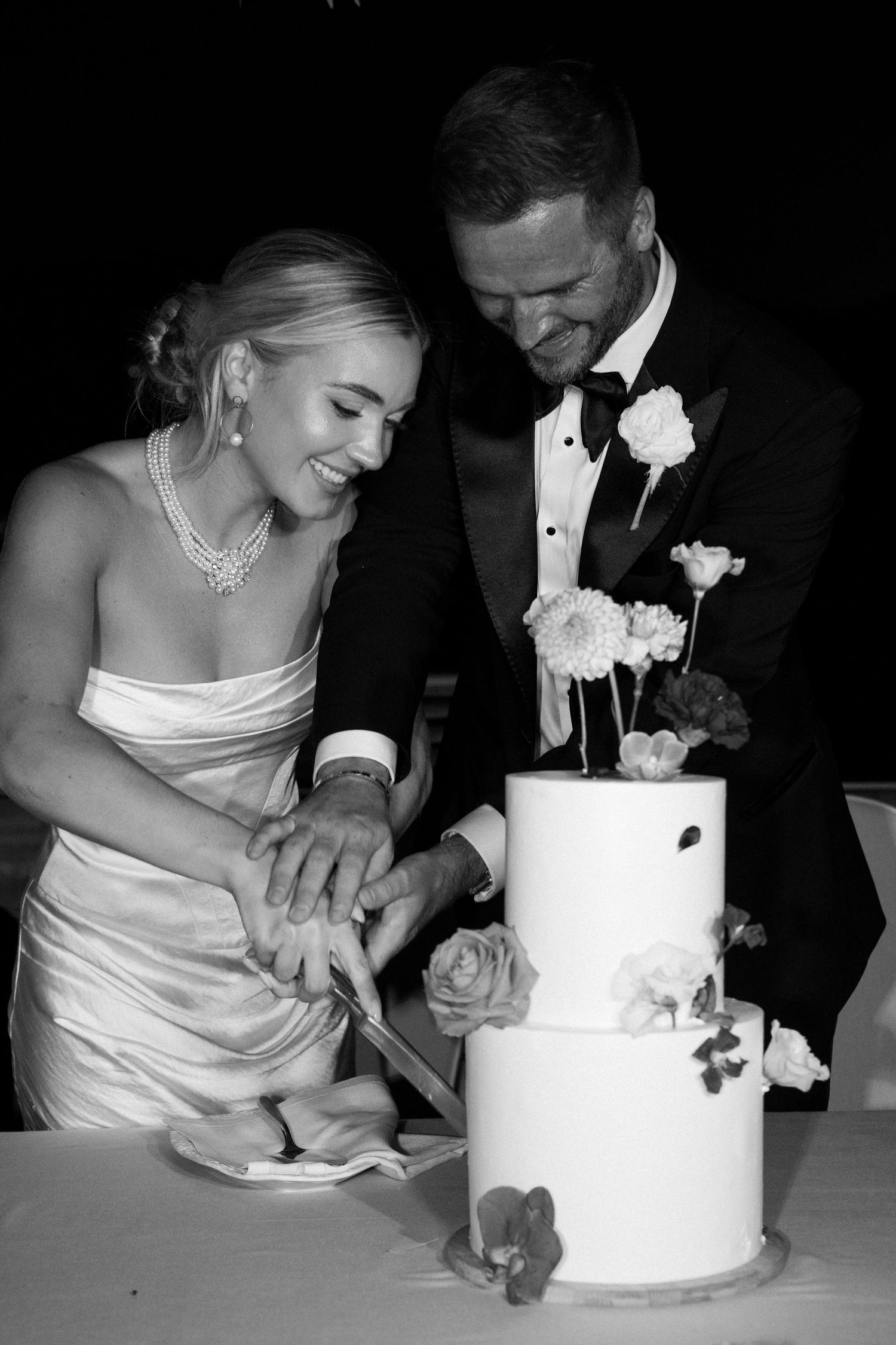 Newlyweds cutting their white tiered wedding cake together in black and white.