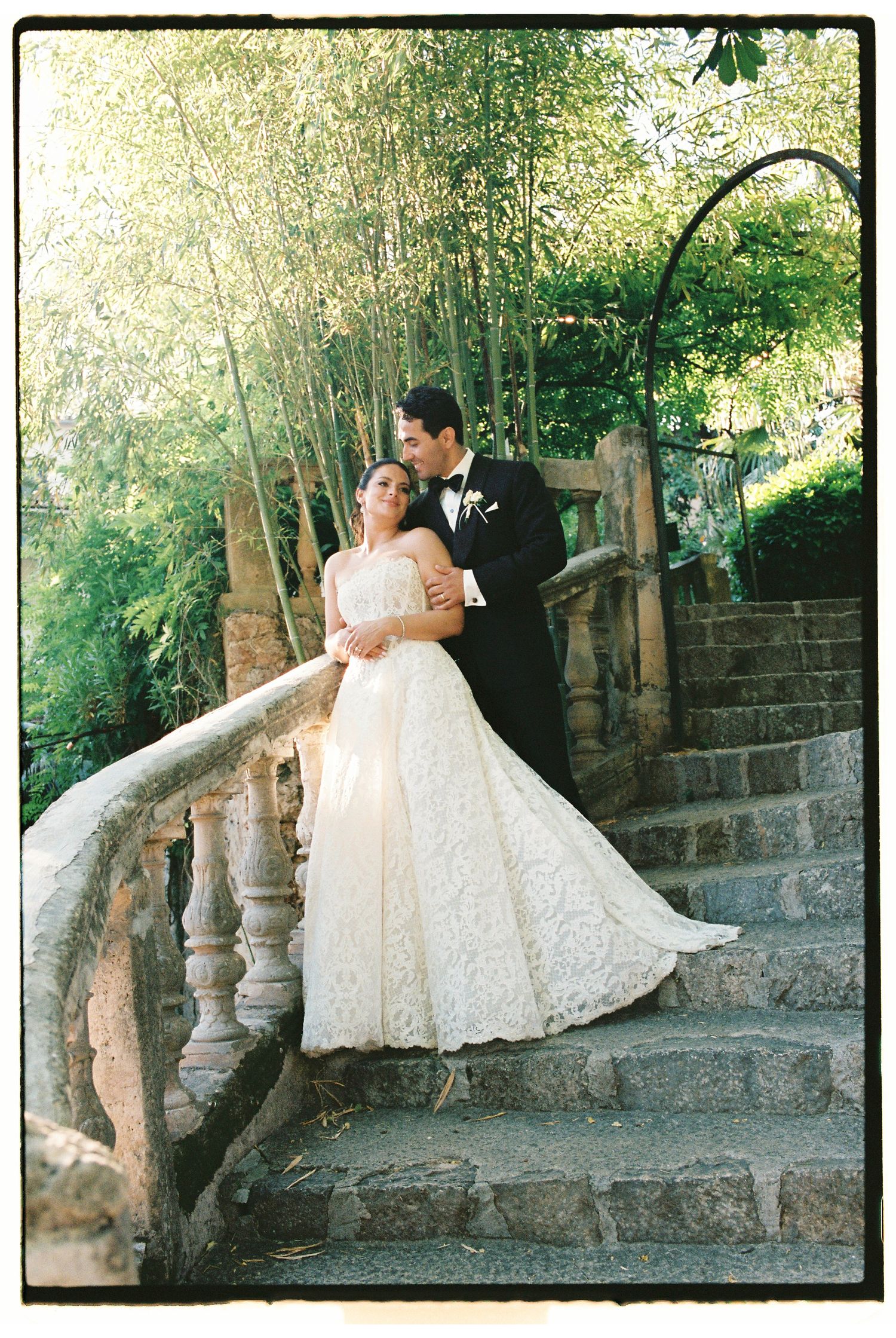 Bride in lace gown and groom in tuxedo pose romantically on stone staircase surrounded by lush greenery.