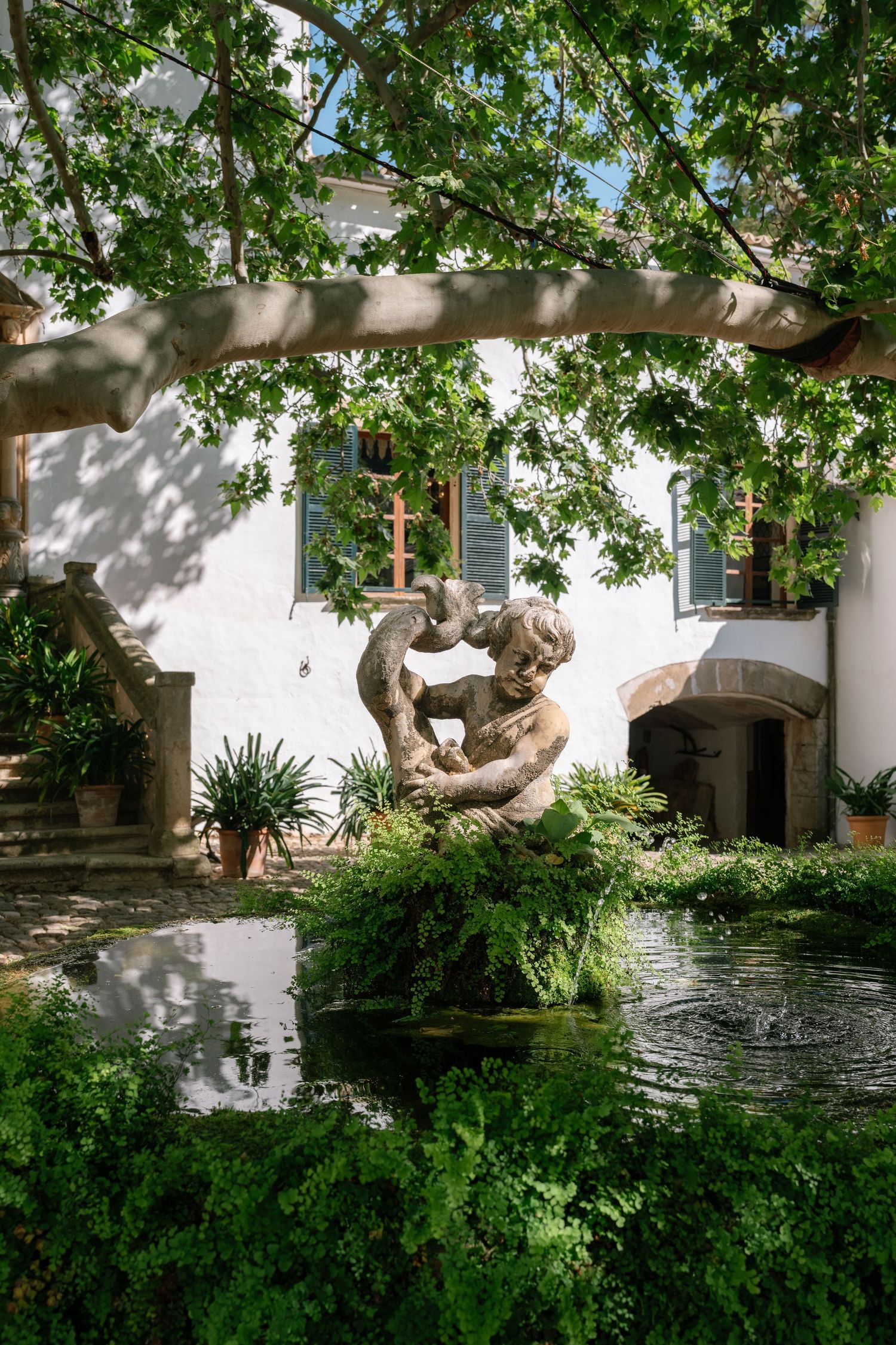 Stone sculpture fountain surrounded by lush greenery and ferns beside a white Mediterranean villa courtyard.