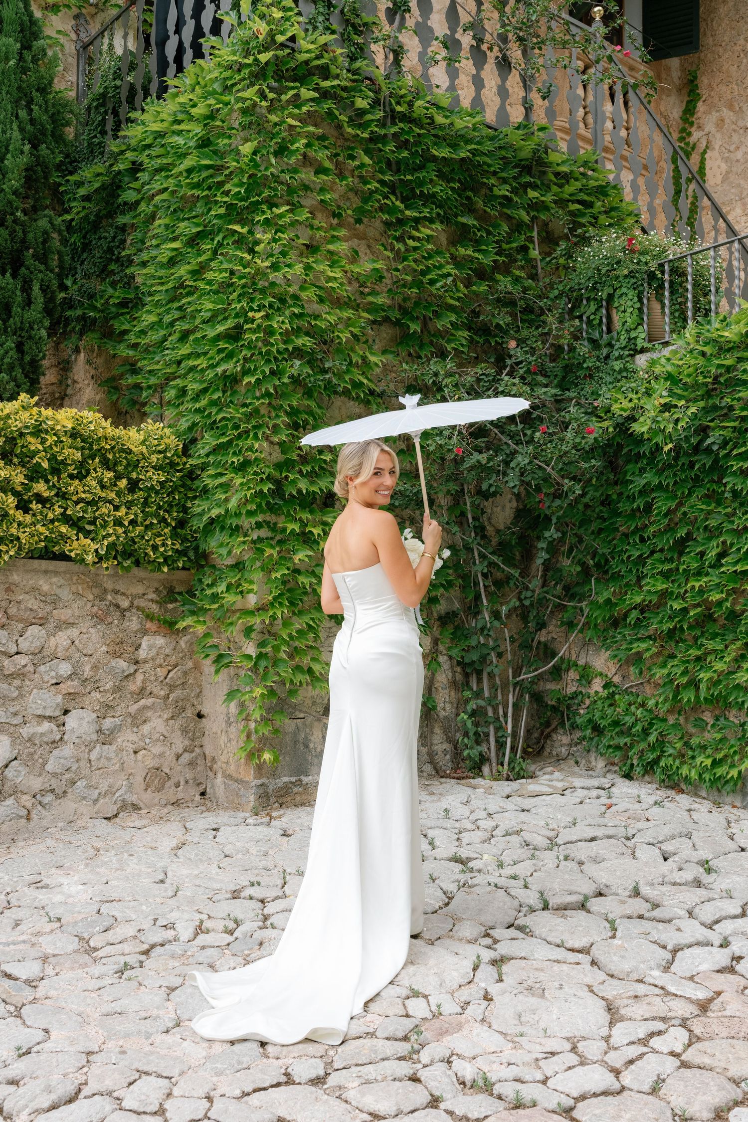 Bride in white strapless gown holding a white parasol umbrella, standing on stone path with ivy-covered wall backdrop.