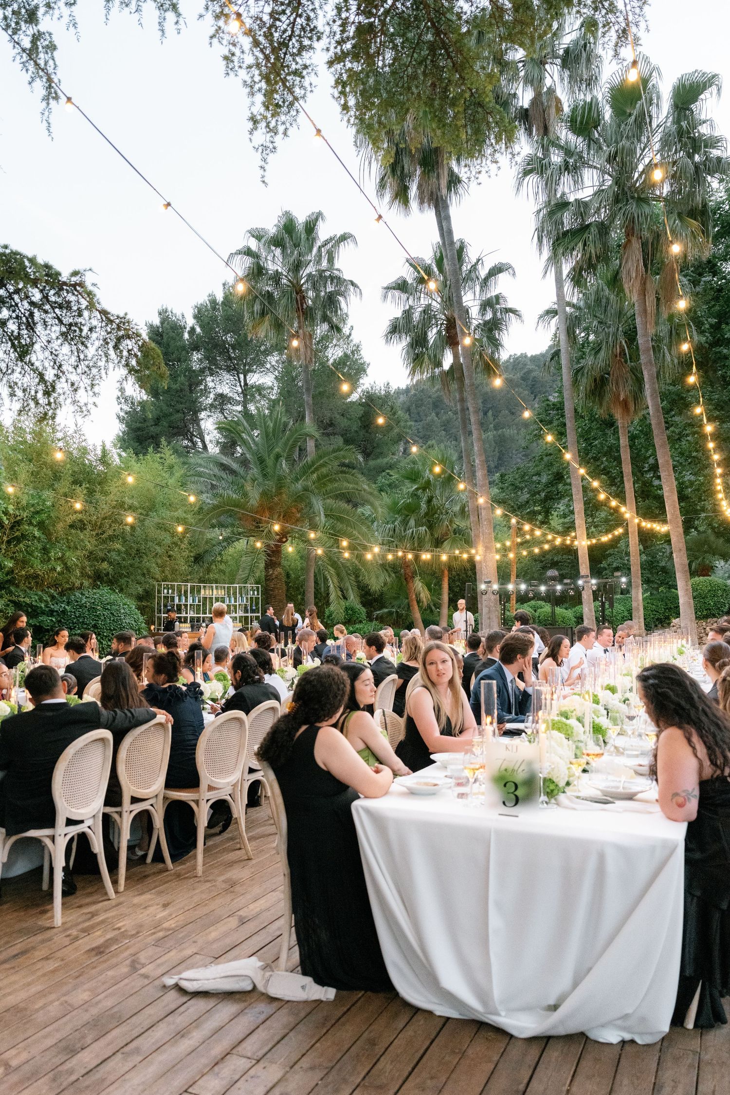 Outdoor wedding reception on wooden deck with string lights, white tablecloths, and palm trees at dusk.