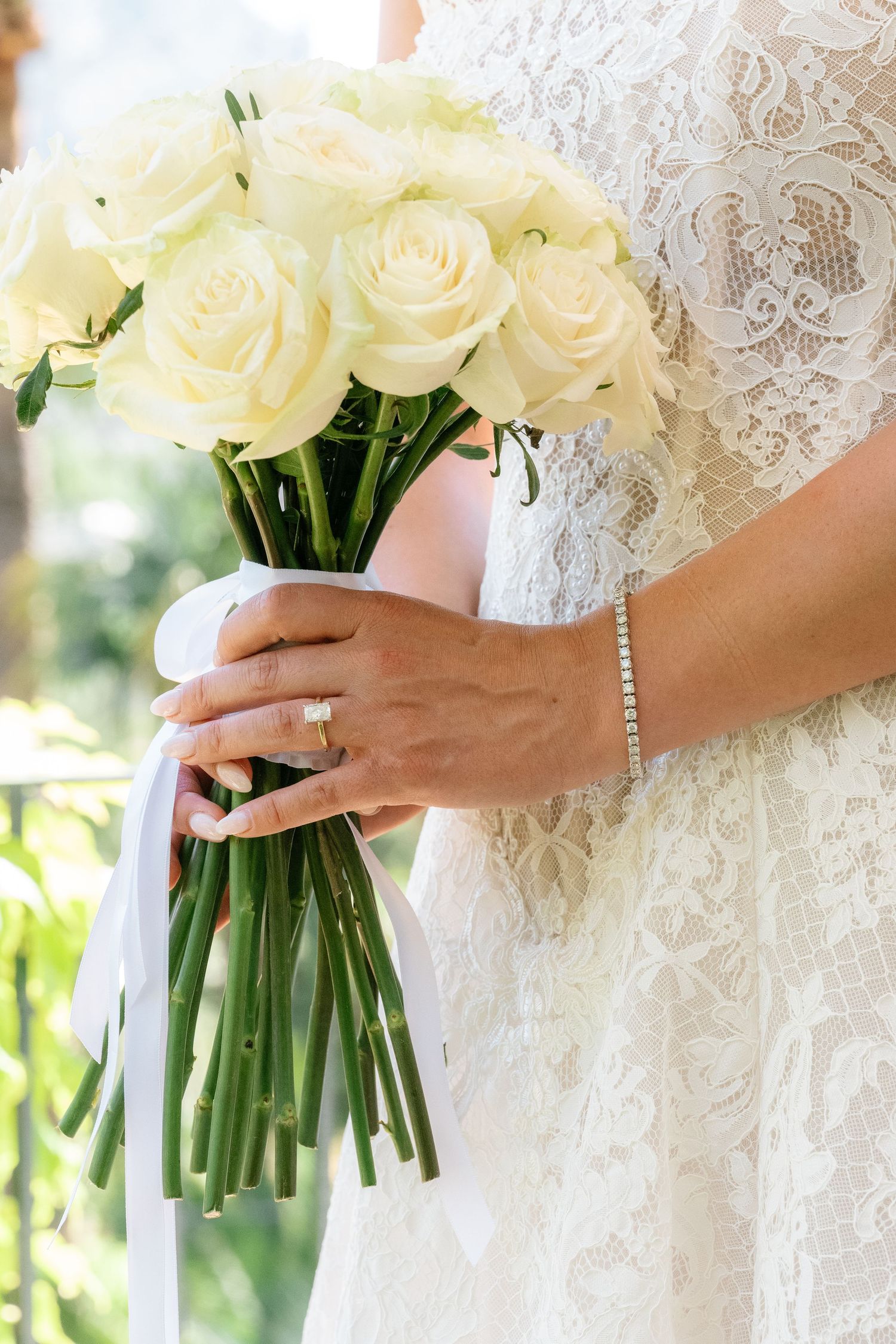 Bride in lace wedding dress holding elegant bouquet of white roses tied with ribbon, wearing diamond ring and bracelet.