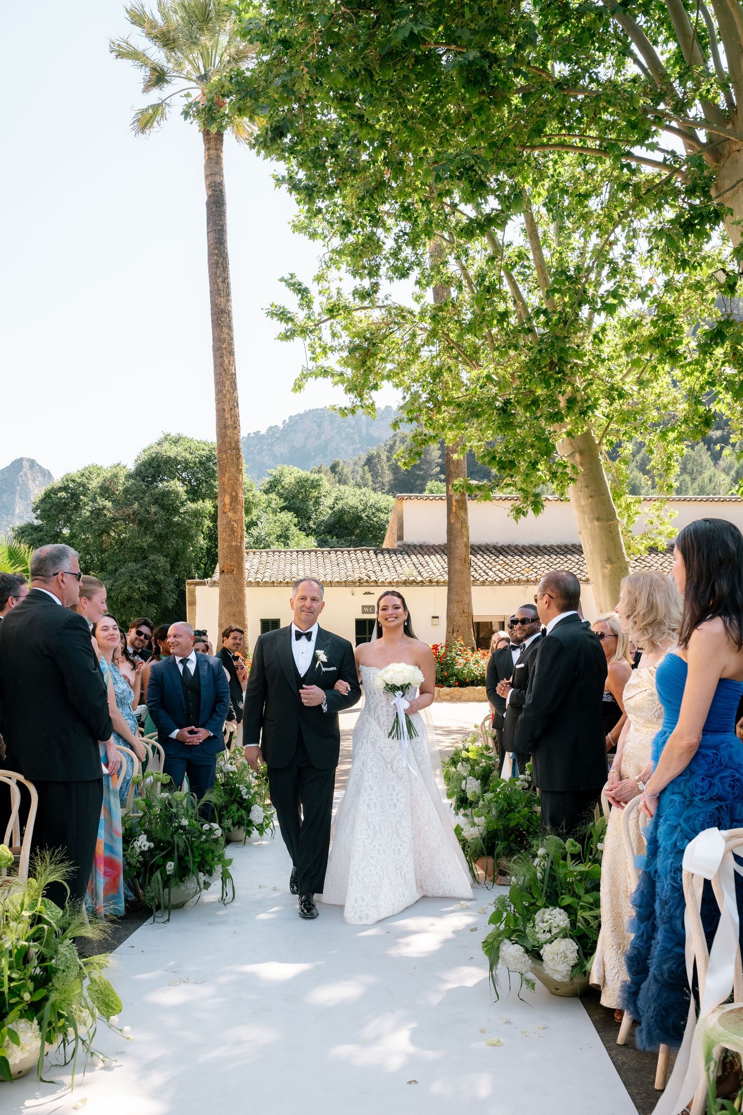 Bride walks down outdoor aisle with escort at sunny garden wedding ceremony, mountains and palm trees in background.
