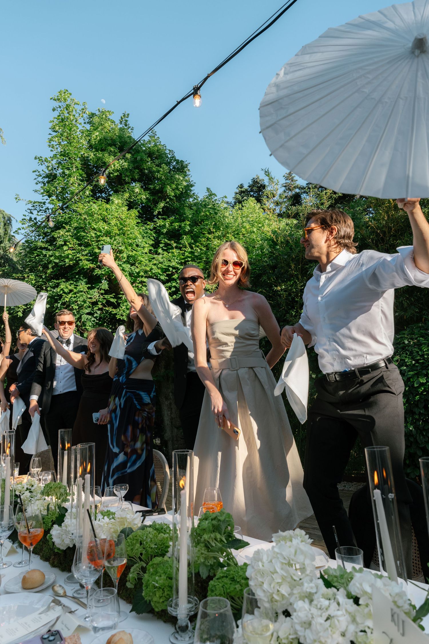 Guests celebrate at an outdoor wedding reception, waving white parasols beside a floral-decorated table under string lights.