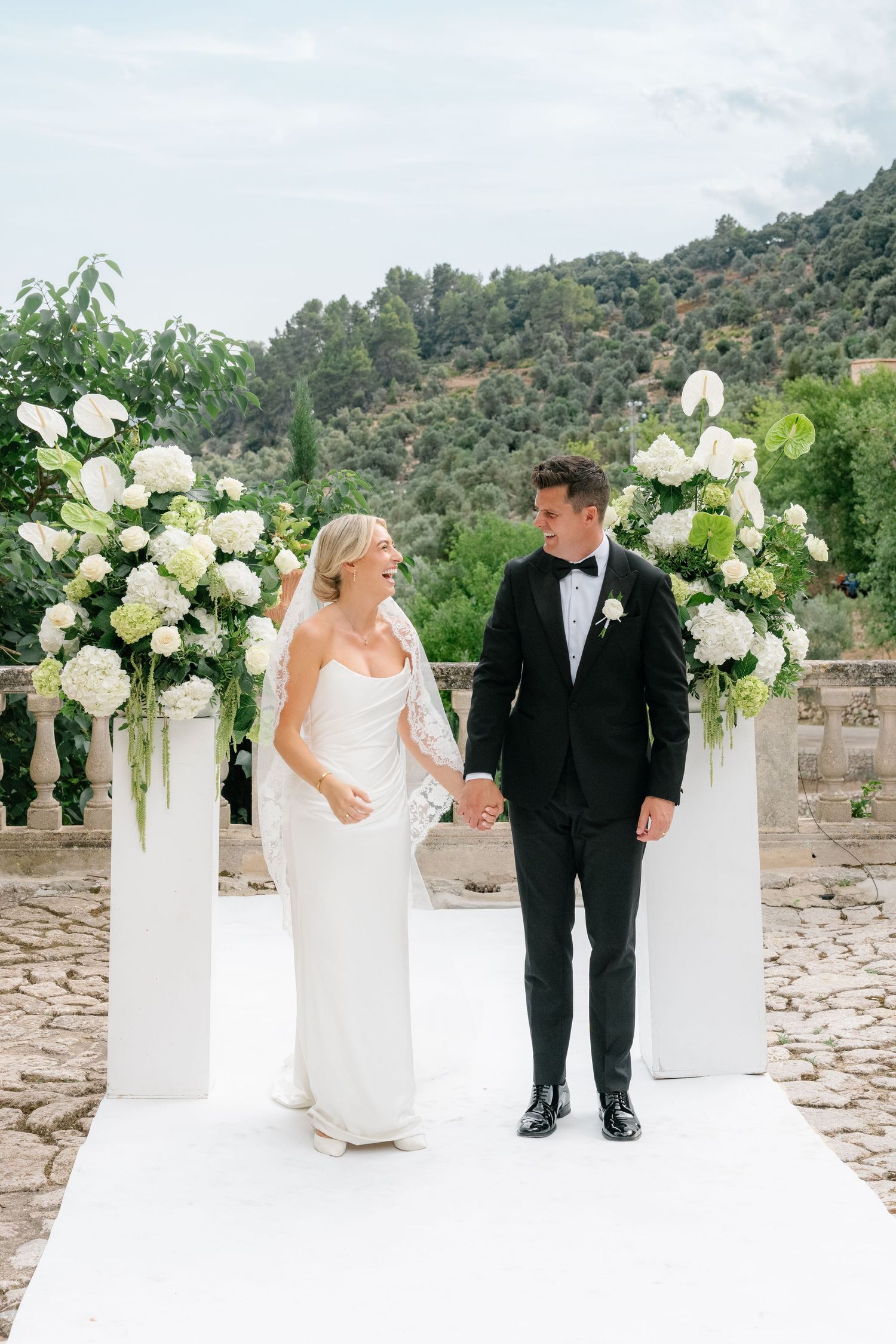 Bride and groom hold hands at outdoor wedding ceremony with white floral arrangements and lush green hillside backdrop.