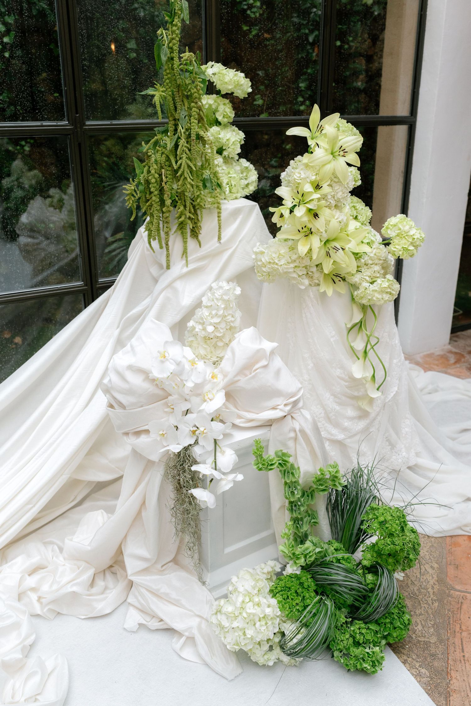 Elegant white wedding dress with flowing fabric and green floral arrangements displayed in a window setting.