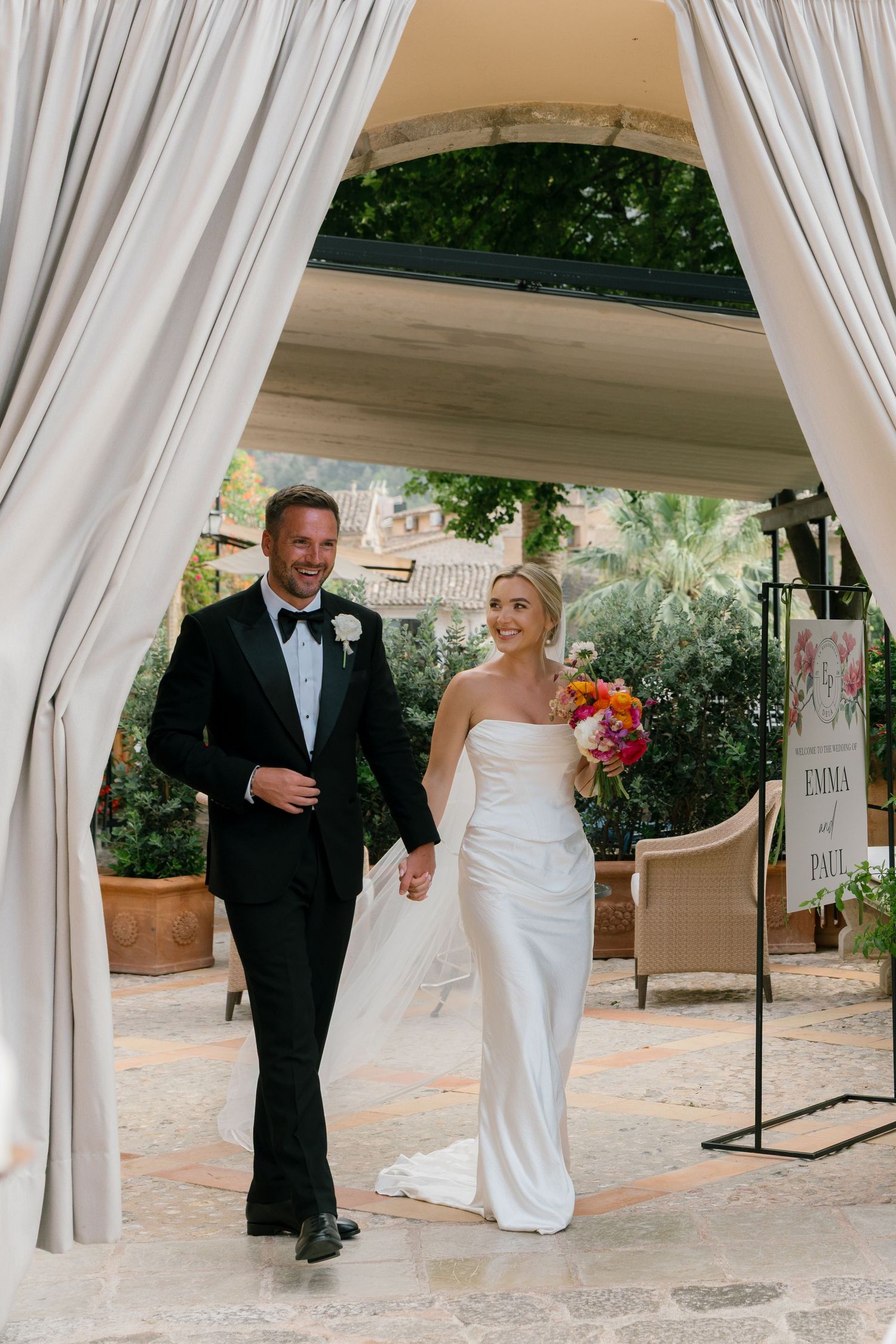 Wedding couple walking through an elegant covered walkway with white drapes and stone architecture.