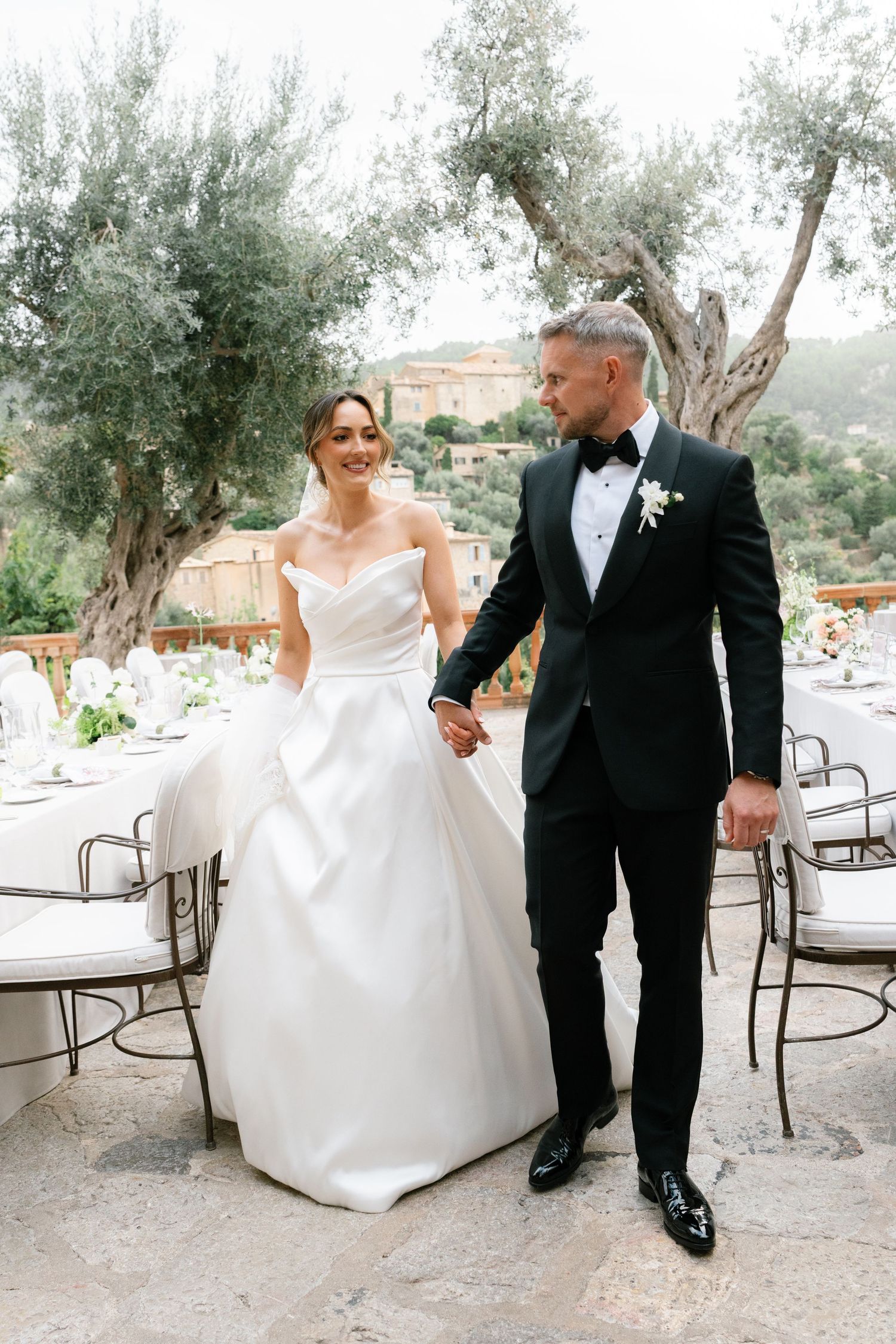 Elegant outdoor wedding reception setup with white tables and chairs under olive trees at a Mediterranean venue.