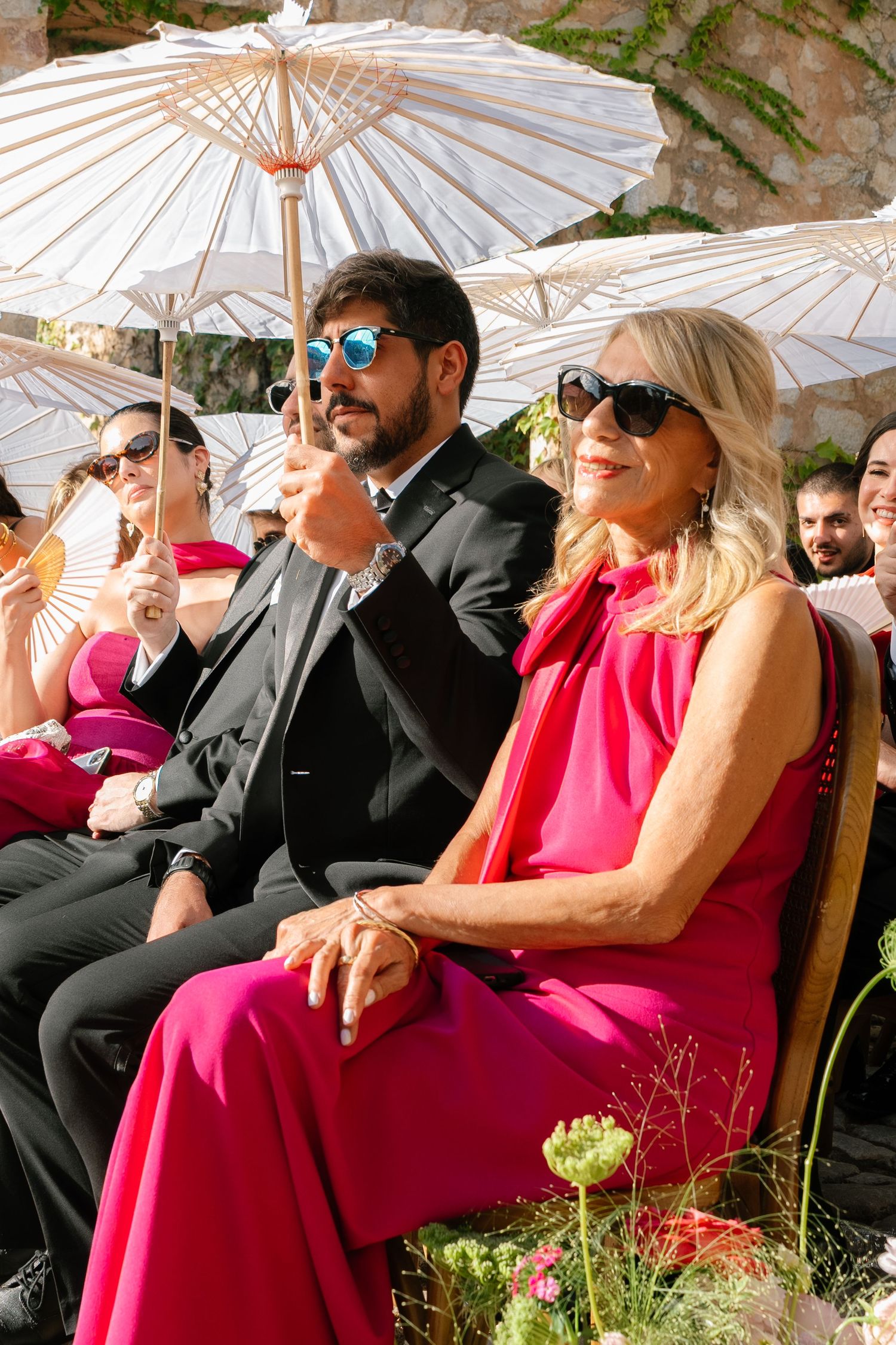 Wedding guests relax under white parasols at an outdoor ceremony with bright pink accents.