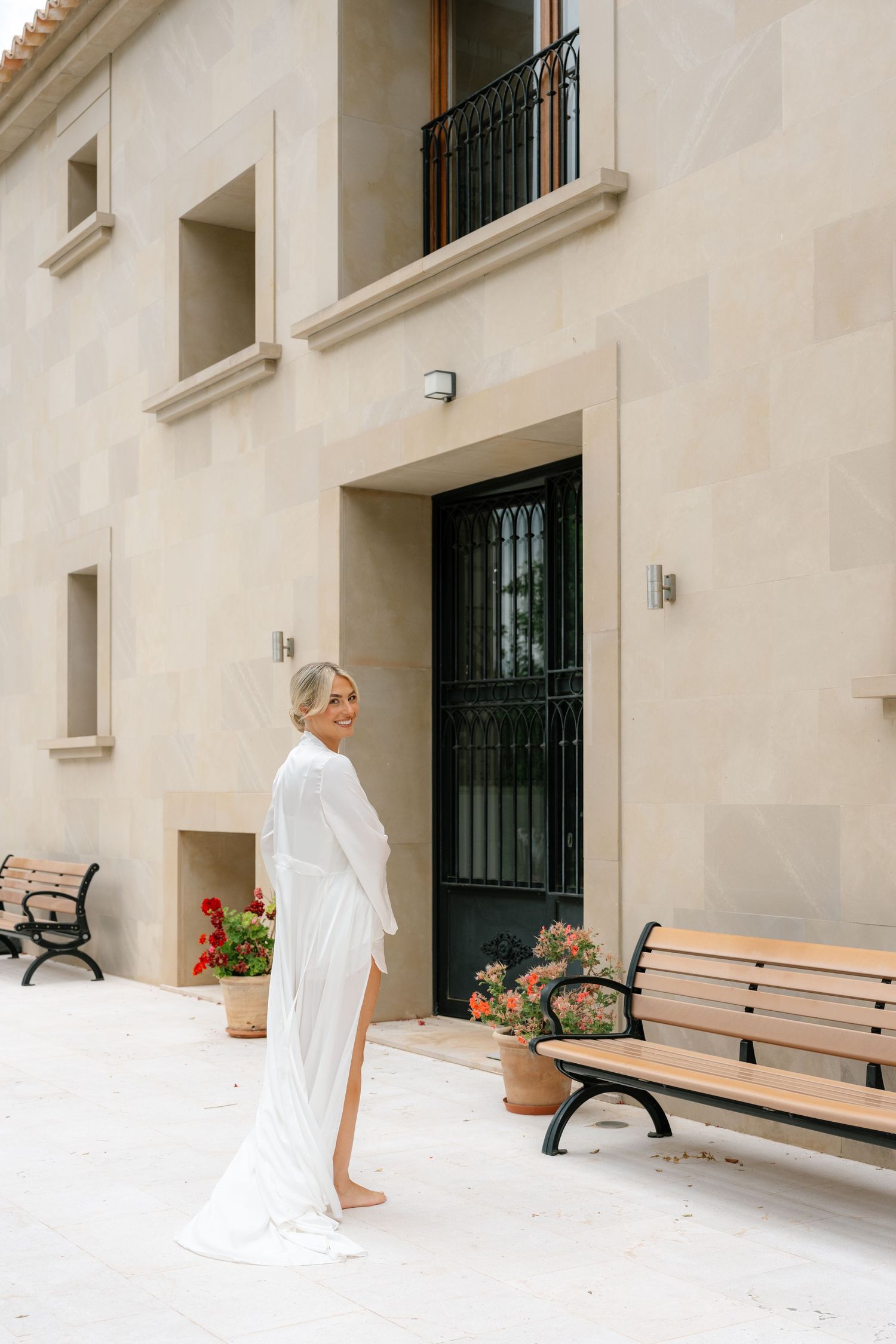 Bride in elegant white long-sleeve gown with slit stands barefoot outside a beige stone building with benches and flowers.