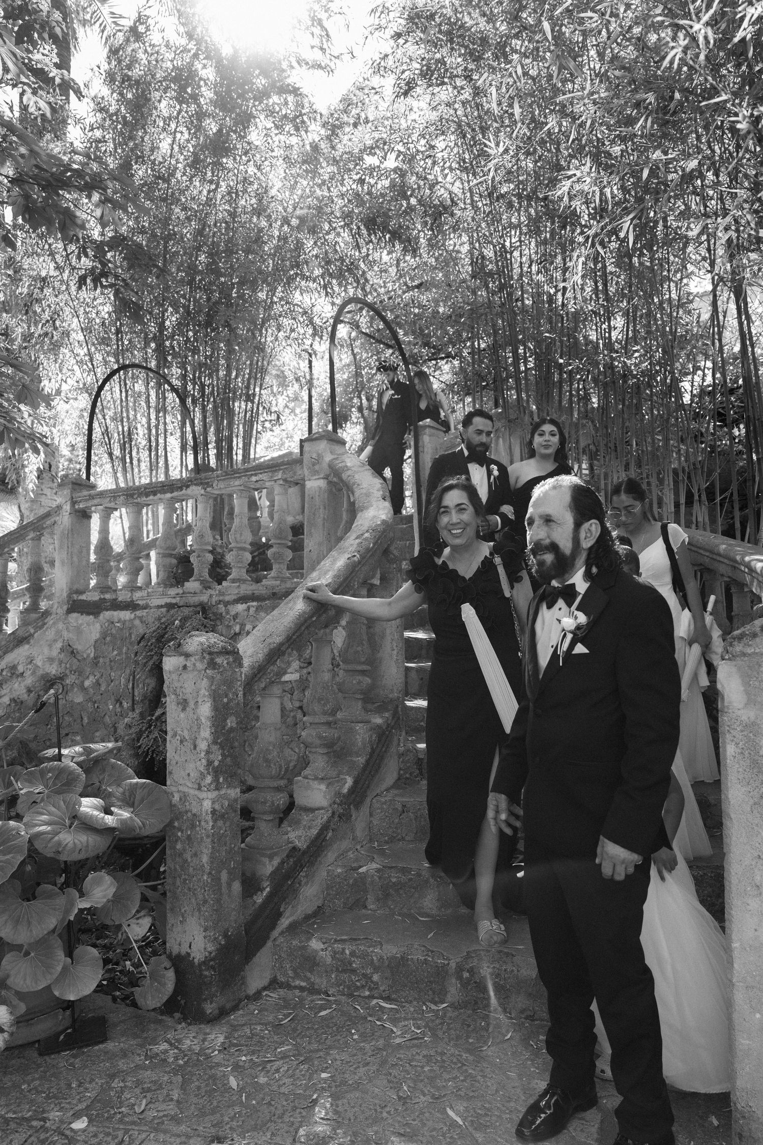Black and white photo of a groom in a tuxedo with wedding guests on stone steps surrounded by bamboo trees.