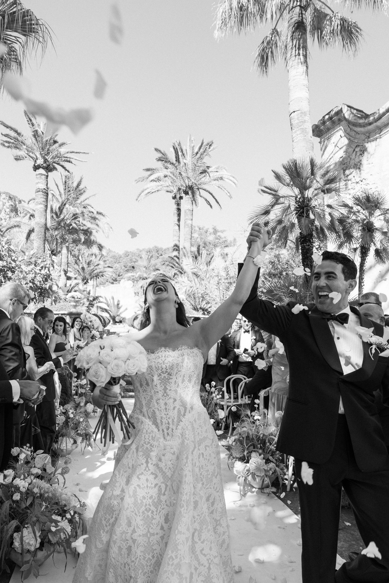 Bride and groom celebrate joyfully walking down outdoor aisle lined with flowers and palm trees at elegant wedding ceremony.
