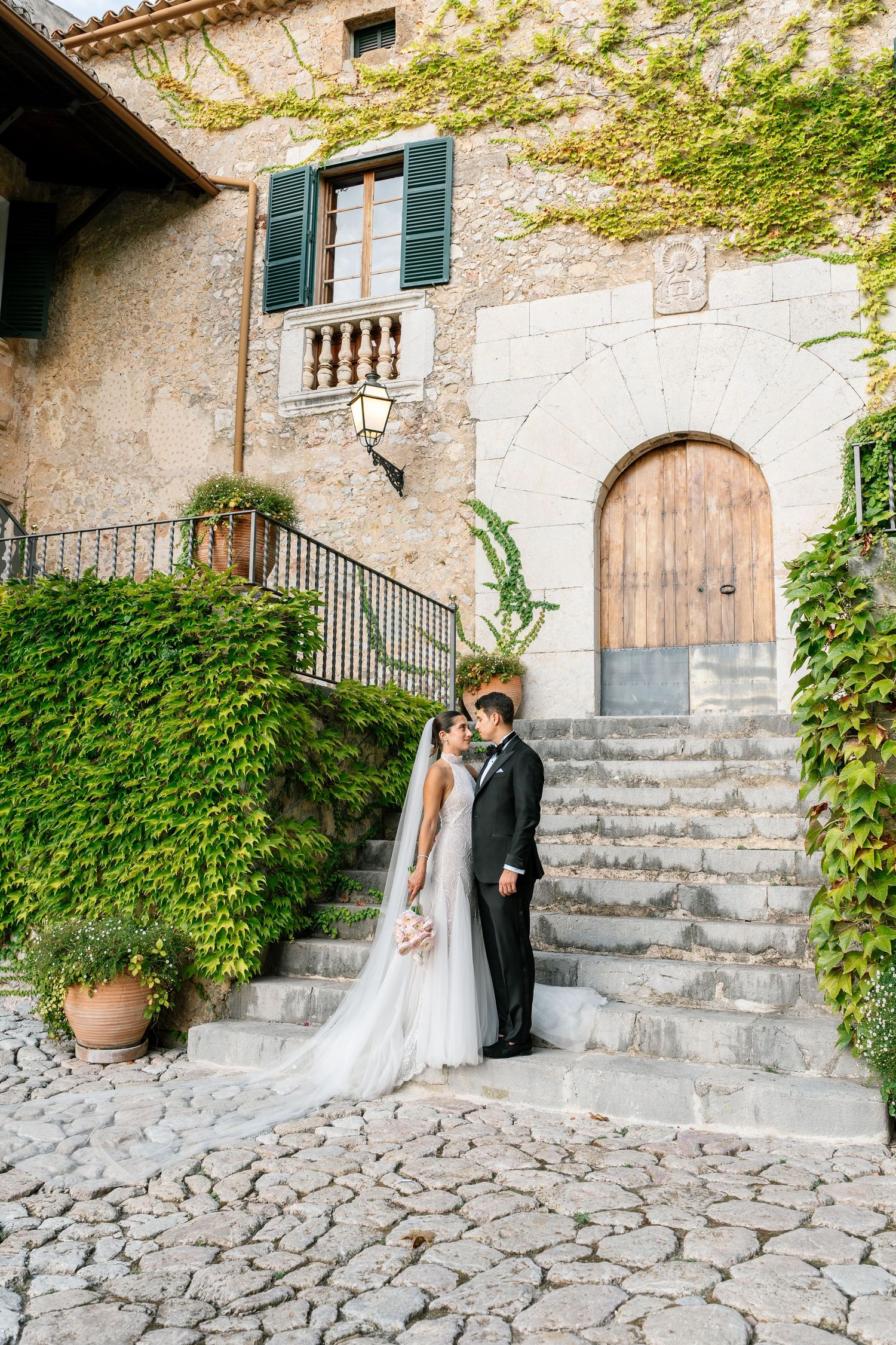 Wedding couple stands on cobblestone steps of a Mediterranean villa with green ivy.