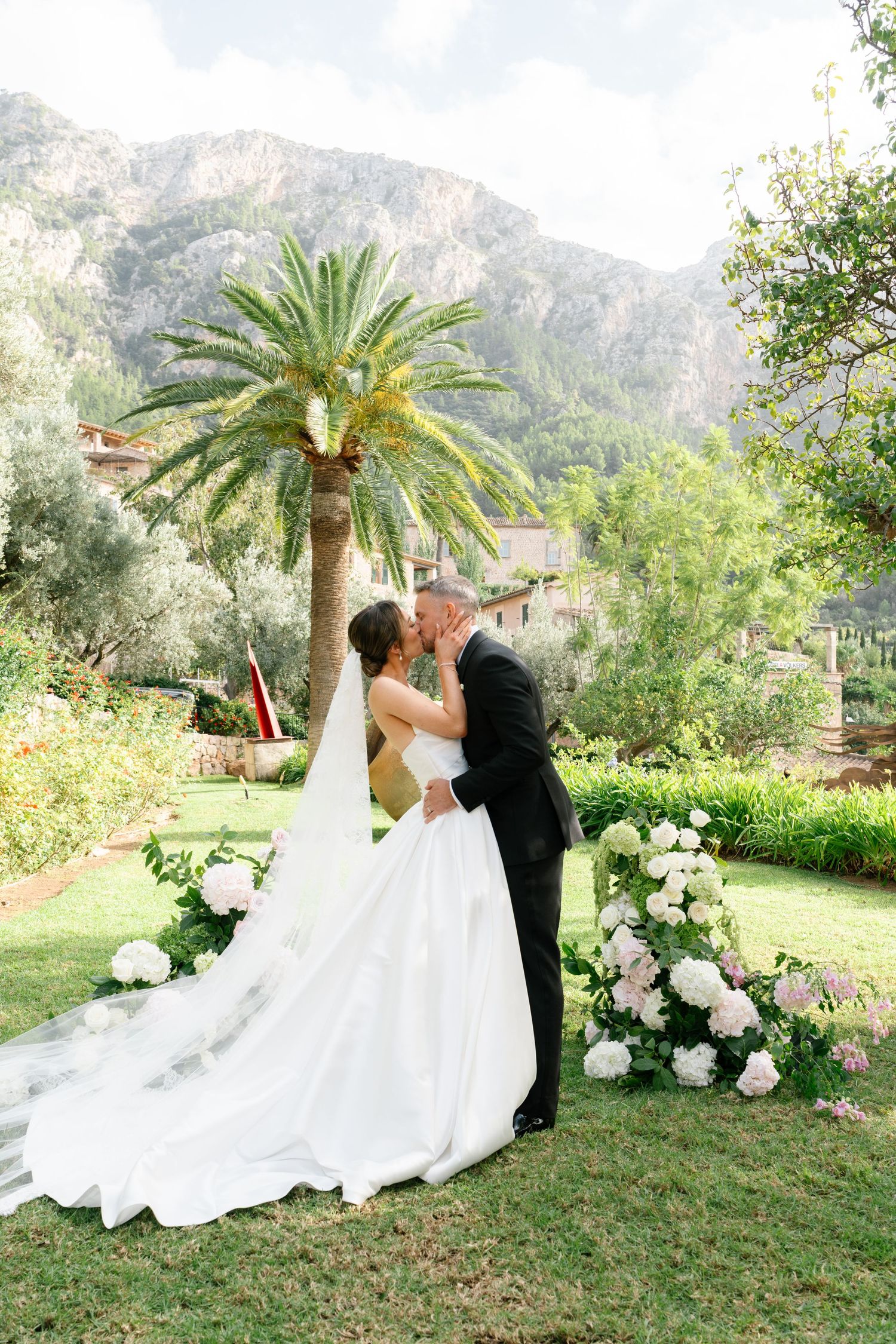 Newlyweds share their first kiss in a garden with palm trees and mountains as backdrop.
