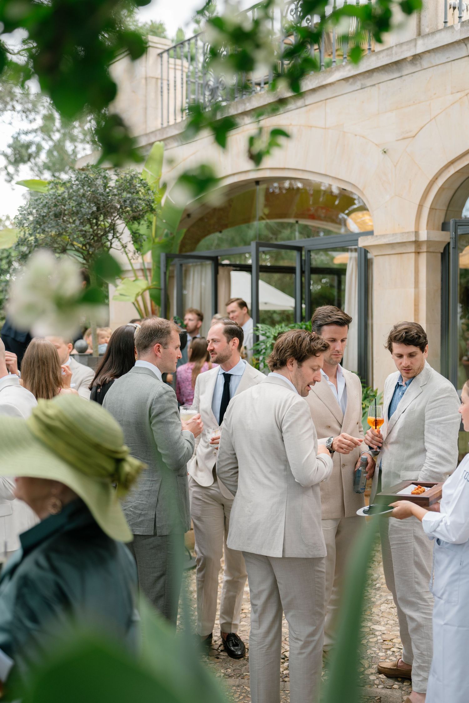 Wedding guests in light colored suits gather for conversation in a lush garden setting.