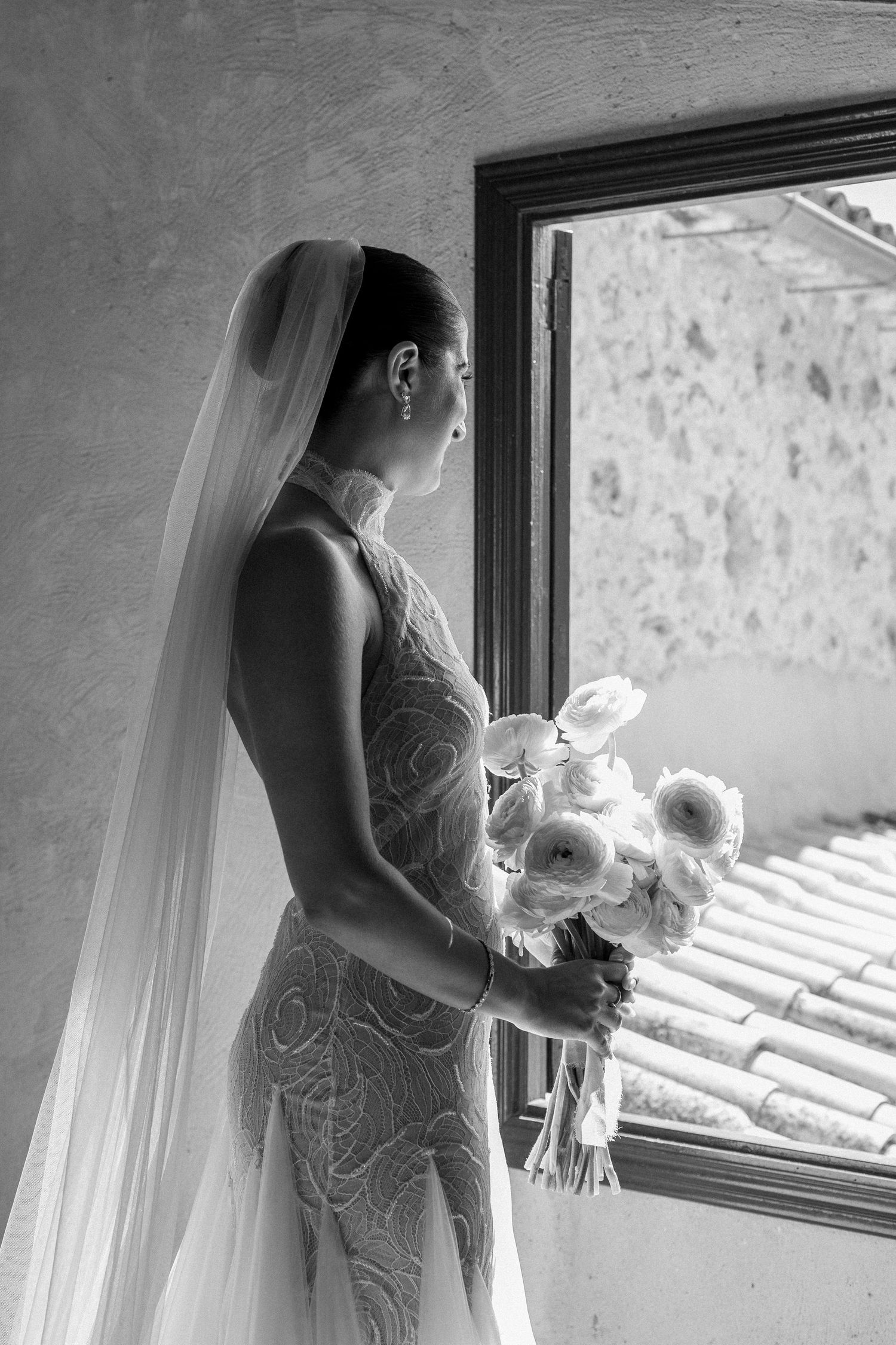 Bride stands by window holding bouquet in black and white sequence.