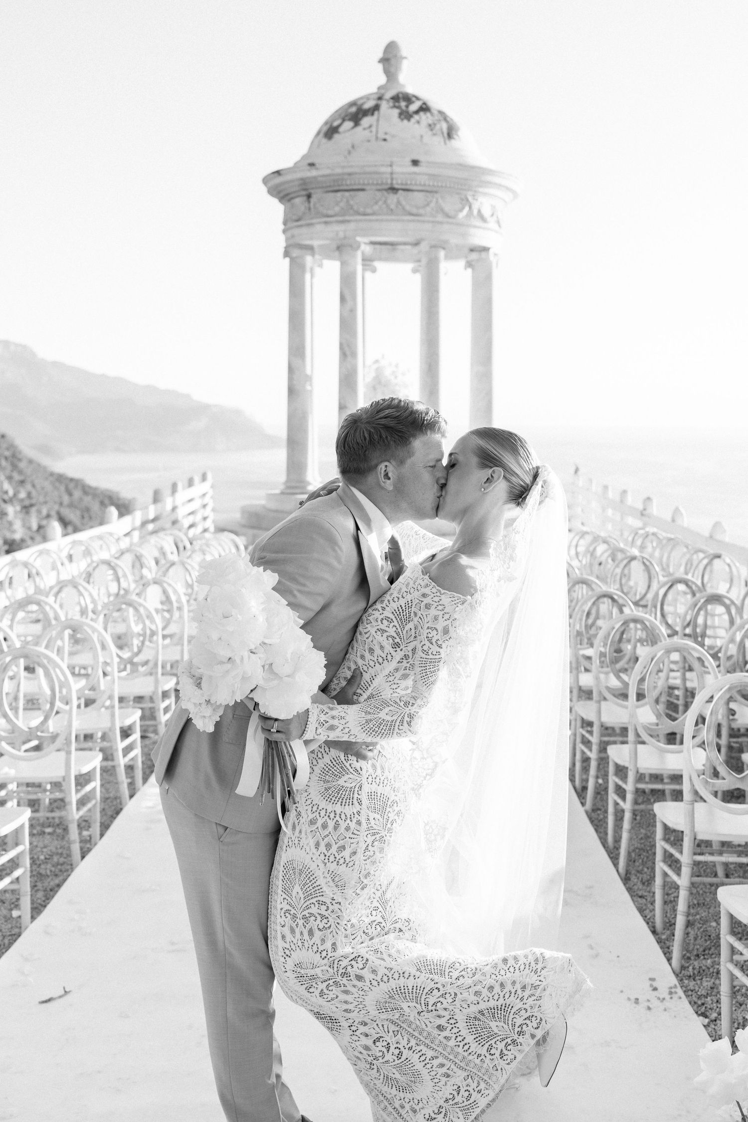 Elegant wedding photoshoot featuring a couple in a classical columned pavilion with white chairs.