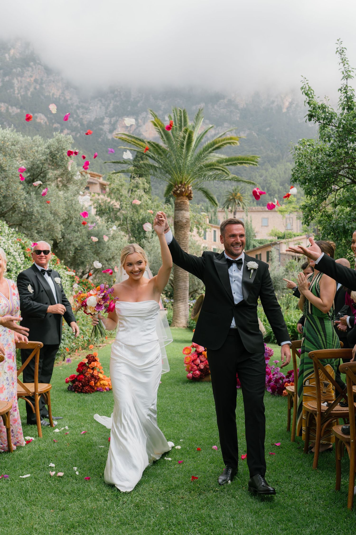 Newlyweds celebrate with raised arms while walking down aisle with palm trees and mountains behind.