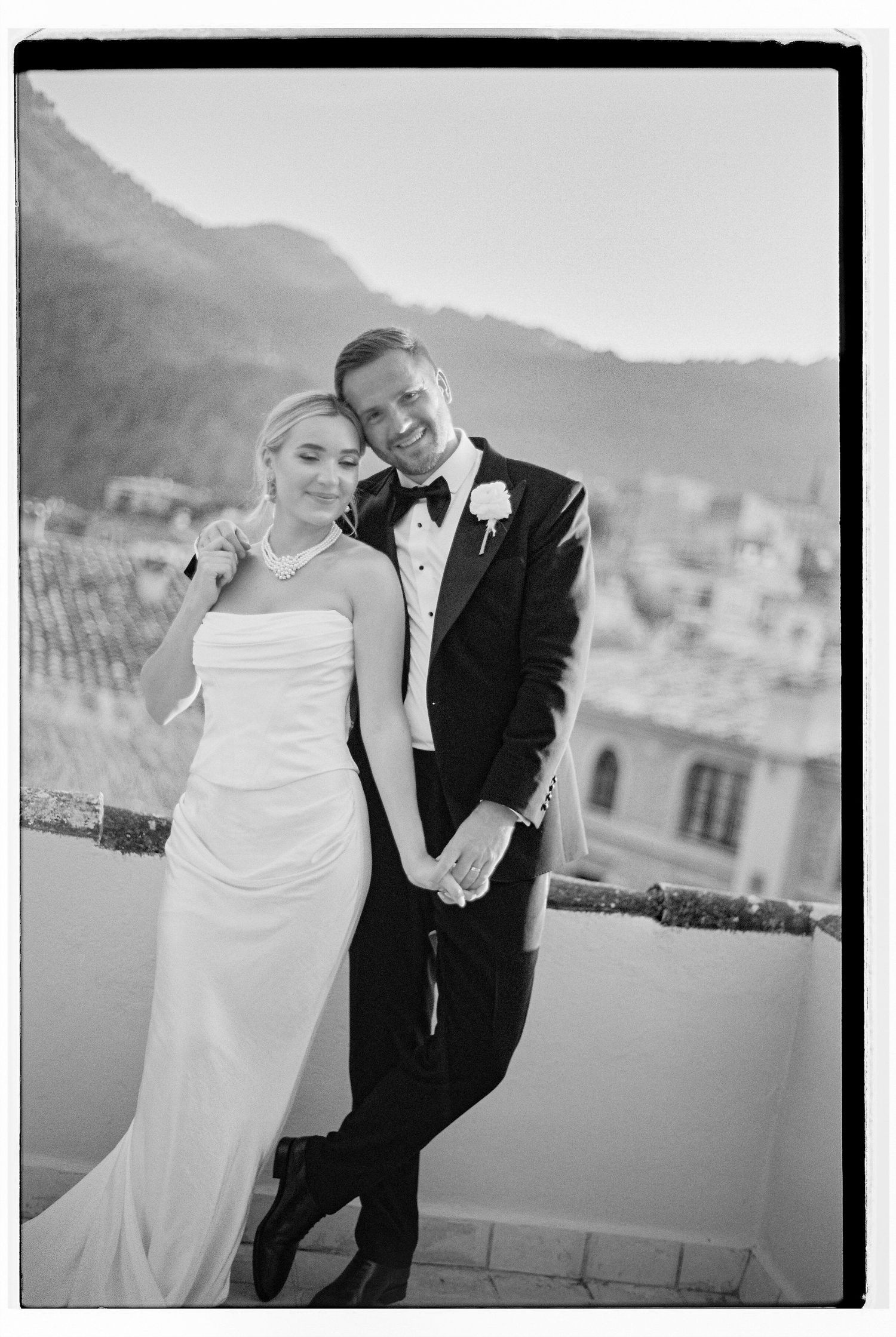 A couple leans against each other while sharing a laugh on a balcony overlooking mountains.