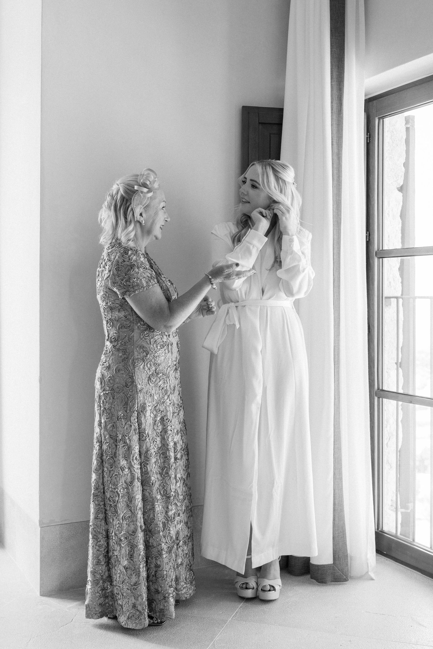 Two women in elegant dresses share a tender moment during wedding preparations.