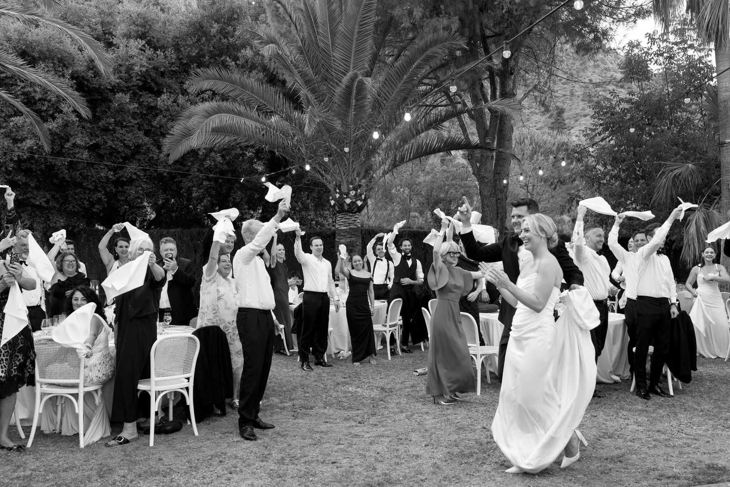 Black and white wedding photo of bride and guests waving white handkerchiefs outdoors under string lights and palm trees.