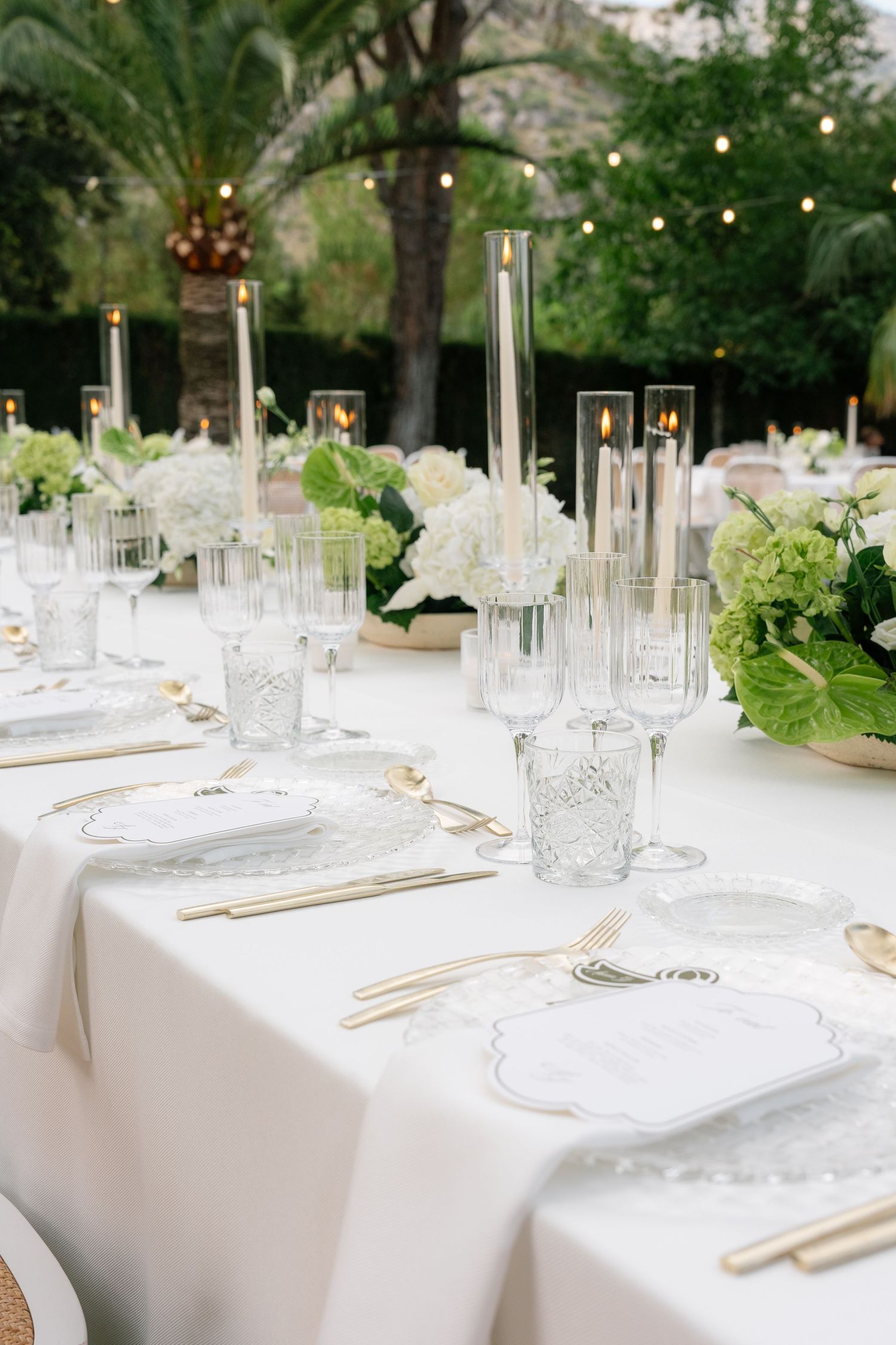 Elegant outdoor wedding table setting with white linens, gold cutlery, crystal glassware, green hydrangeas, and tall candle holders.