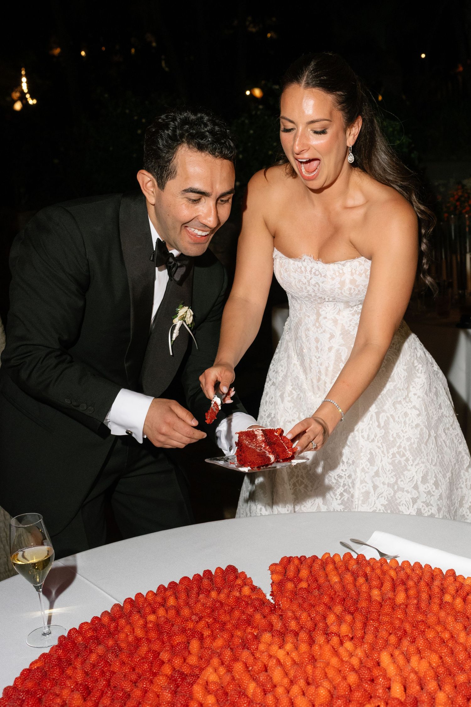 Bride and groom smiling while cutting a red wedding cake surrounded by fresh raspberries at their elegant evening reception.