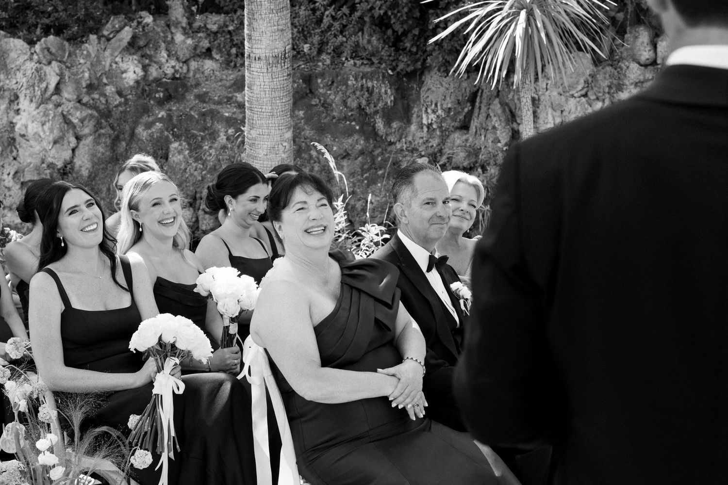 Black and white photo of smiling bridesmaids in dark dresses holding bouquets at an outdoor wedding ceremony.