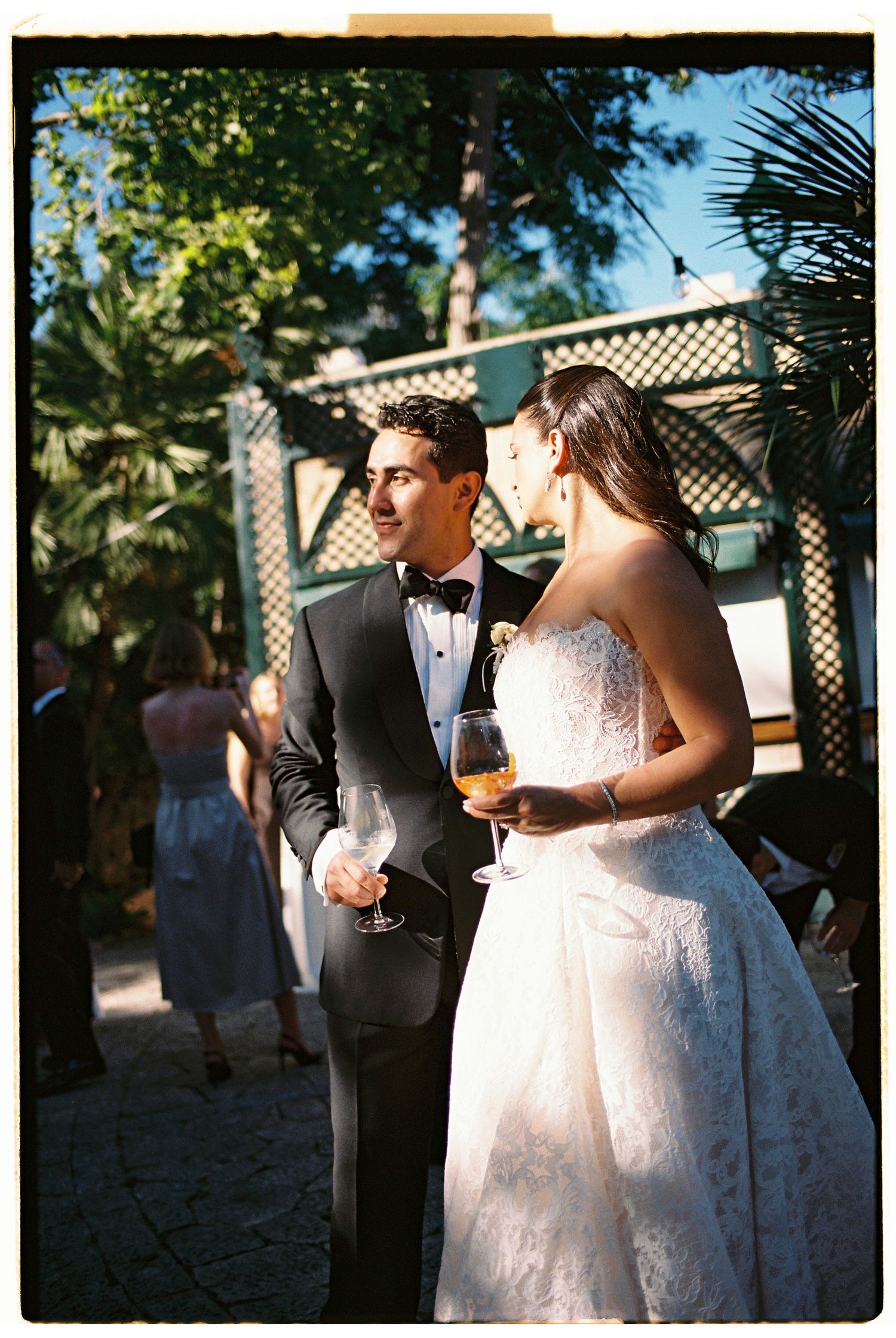 Bride and groom holding champagne glasses at outdoor wedding reception, surrounded by palm trees and string lights.