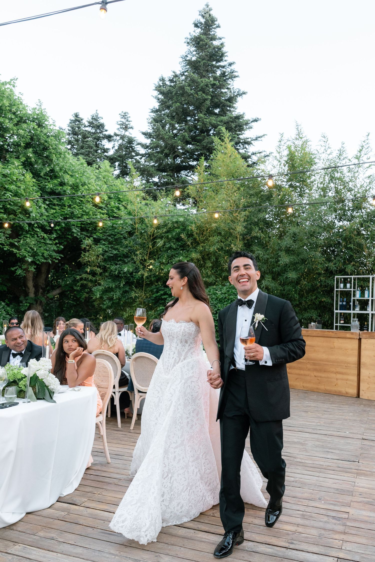 Bride in lace gown and groom in tuxedo walk hand-in-hand at outdoor wedding reception with string lights.