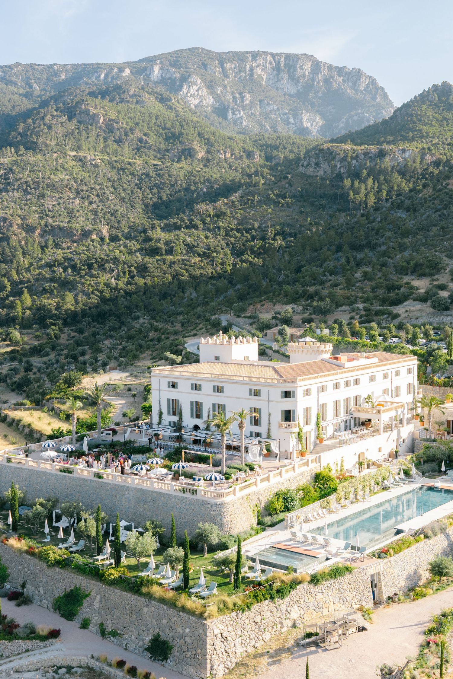 Luxurious white hotel with swimming pool nestled in mountainous terrain of Mallorca with dramatic cliffs in background.