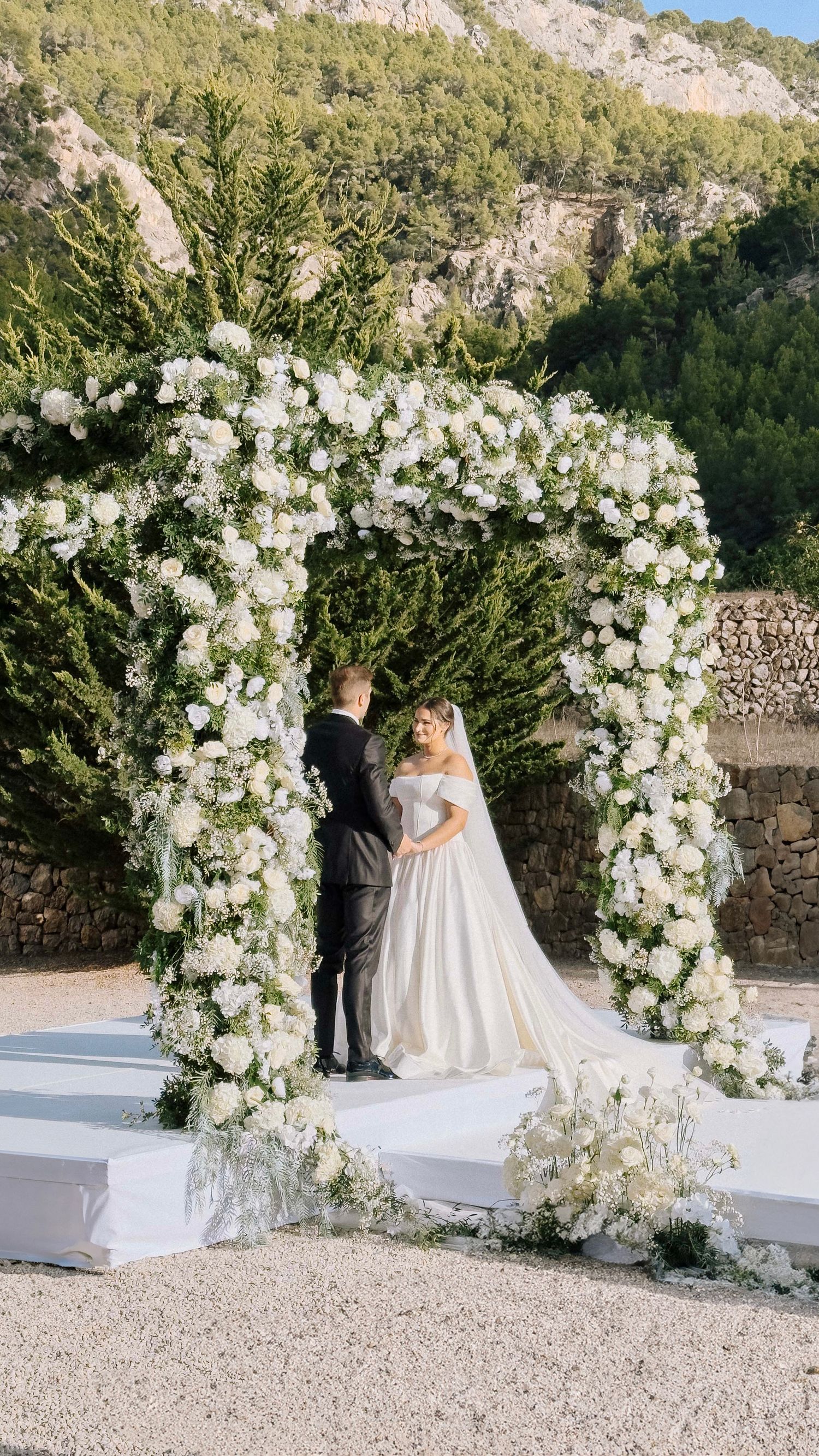 A beautiful outdoor wedding ceremony setup with an elegant white floral arch against a mountainous backdrop.