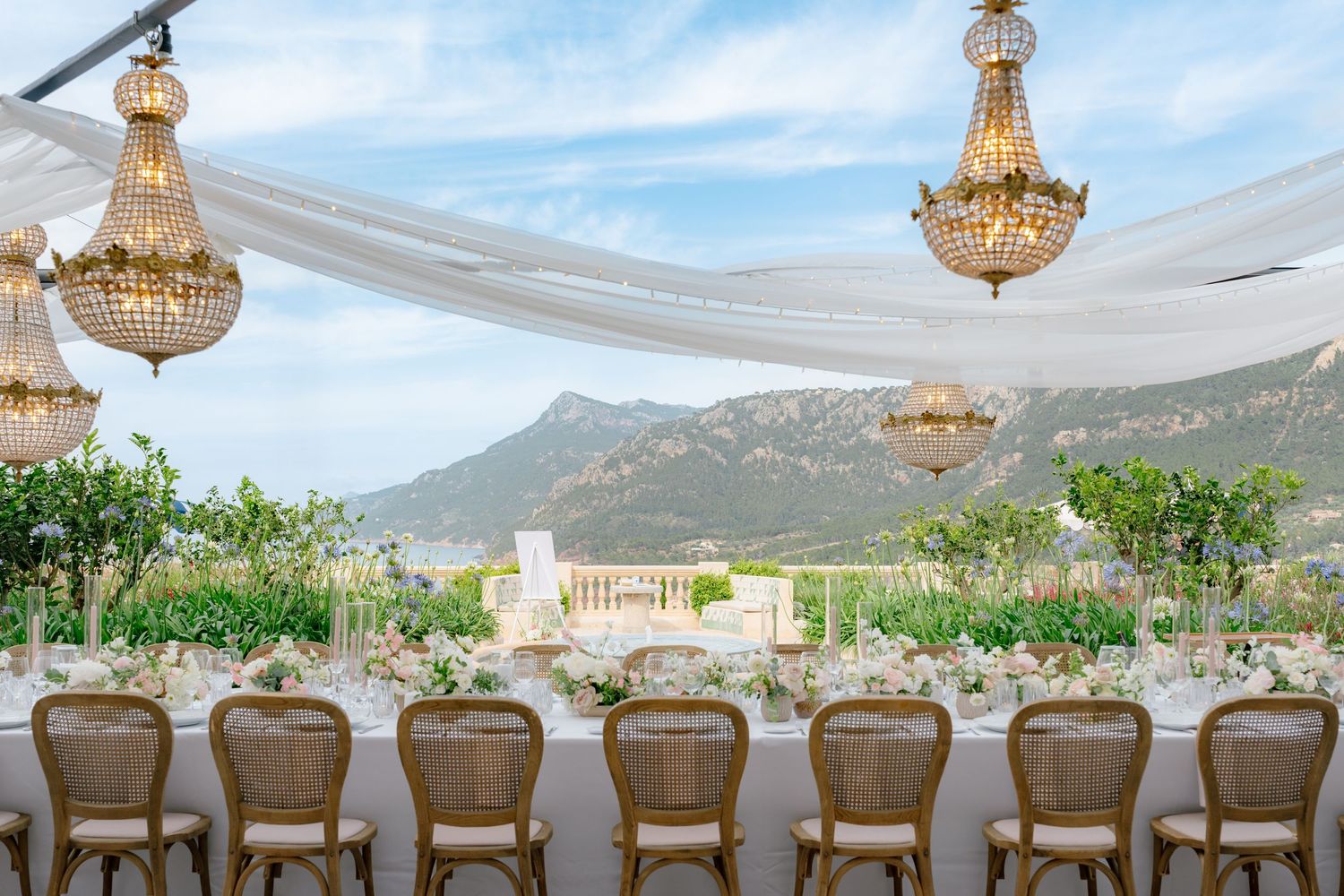 Elegant wedding reception with crystal chandeliers, long dining table, floral arrangements, and stunning mountain backdrop at Son Bunyola