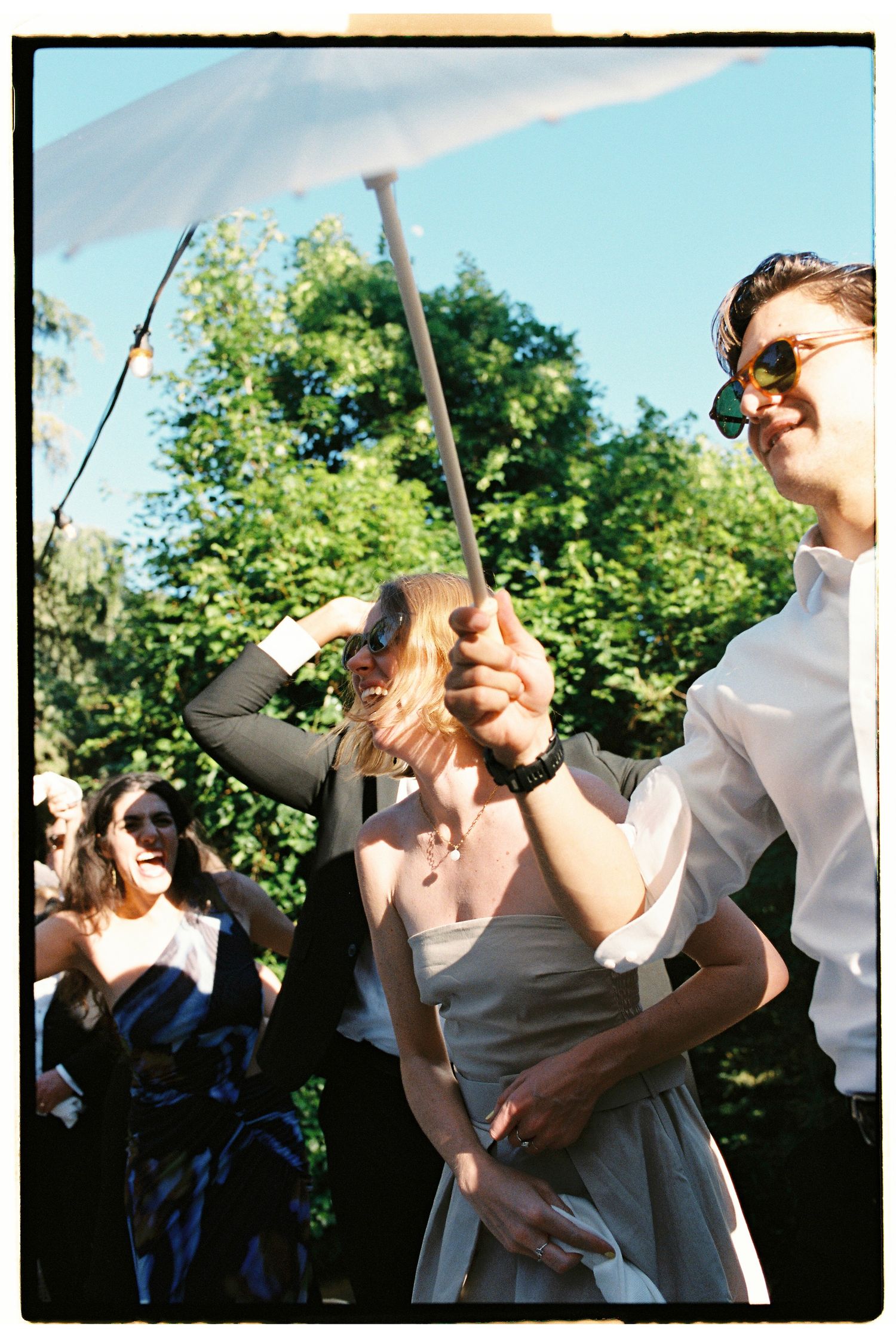 Guests enjoy an outdoor wedding reception on a sunny day, with a woman holding a parasol and a man in sunglasses smiling.